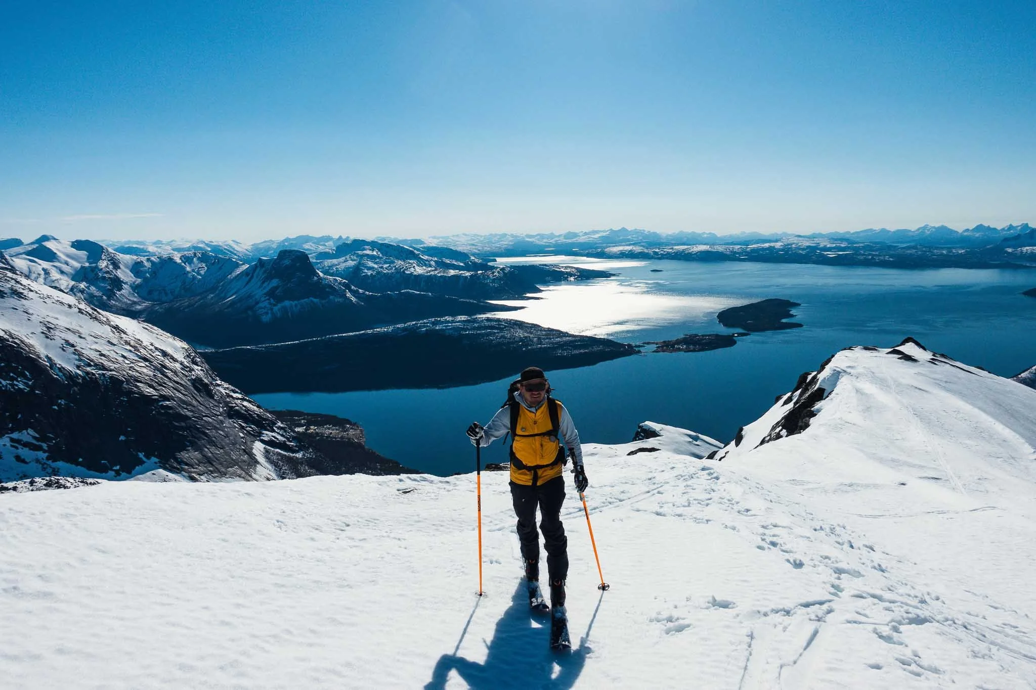 En person som går på ski oppover snødekte fjell med en innsjø i bakgrunnen under en klar blå himmel.