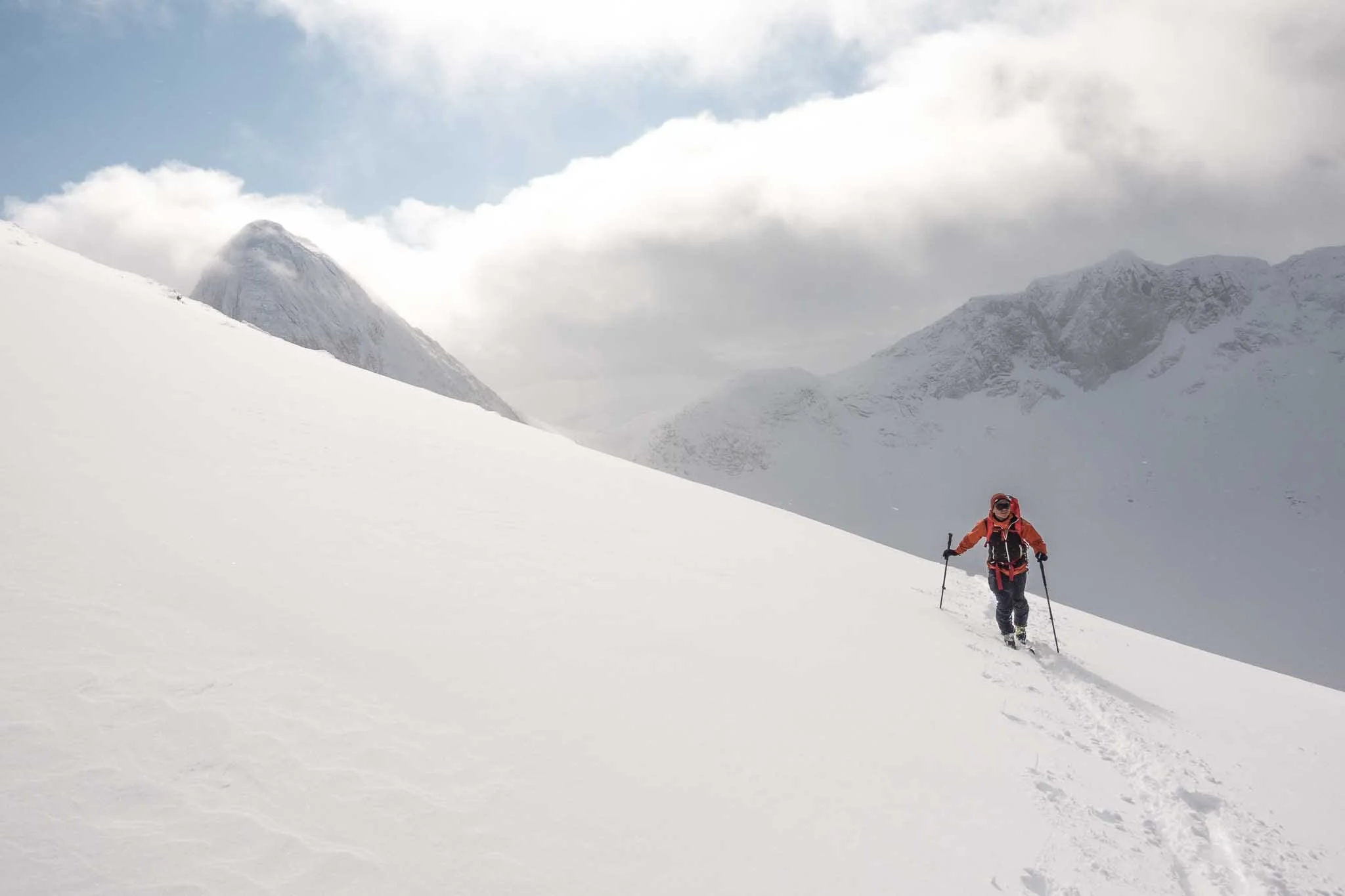 En person går opp en snødekt fjellbakke med ski og staver, omgitt av snødekte fjell og en overskyet himmel.