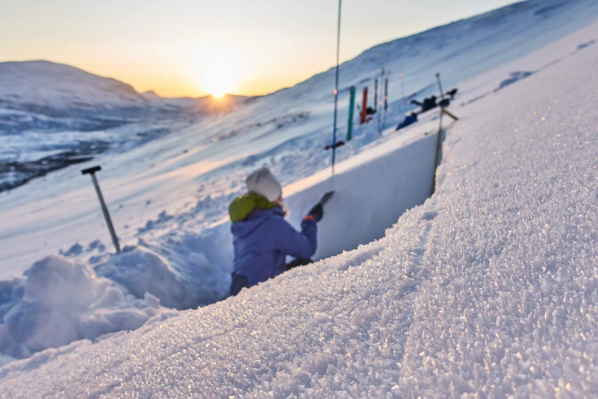 Person som graver i snøen på en fjelltopp med skiutstyr, solnedgang i bakgrunnen.