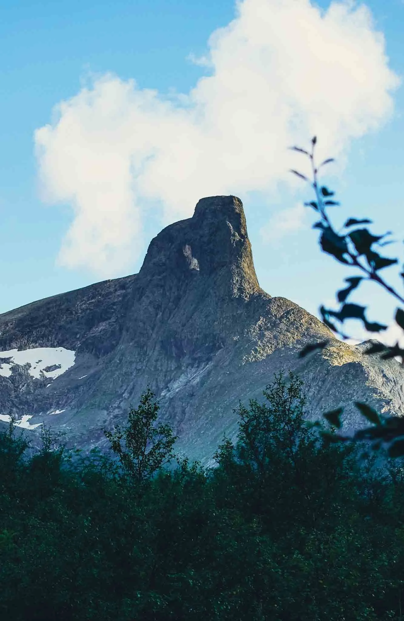 Fjell med en steinformasjon mot en blå himmel med noen skyer og busker i forgrunnen.