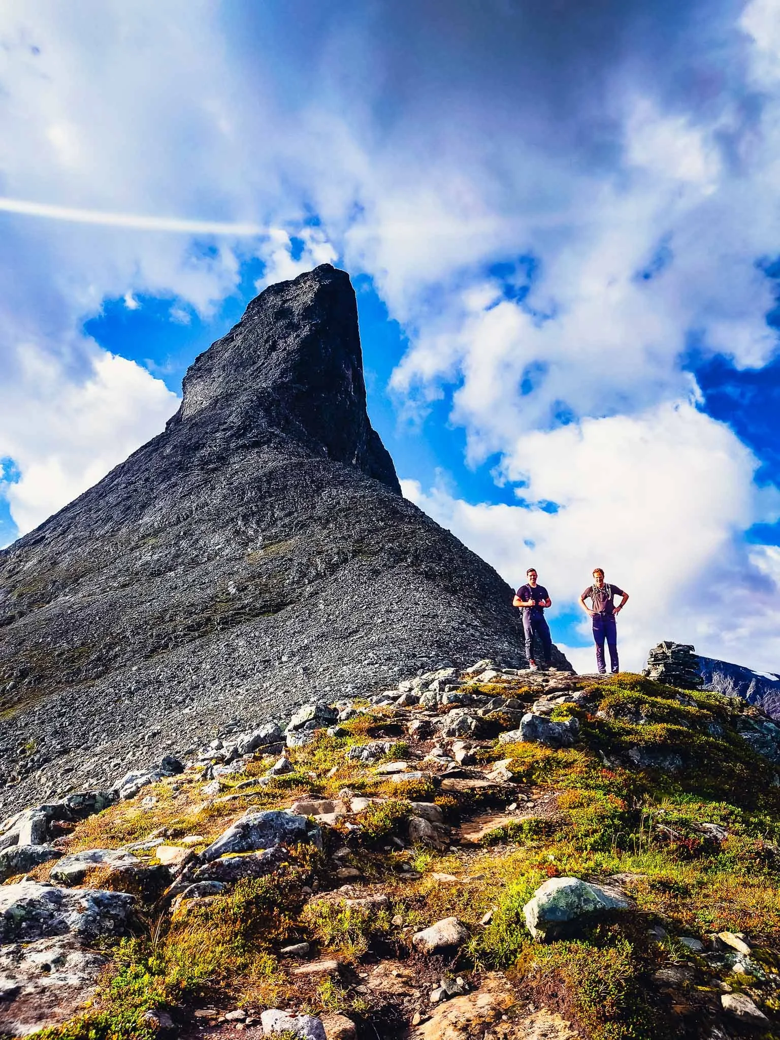 To personer som står på en fjelltopp omgitt av natur, med en stor fjellrygg i bakgrunnen og skyer på himmelen.