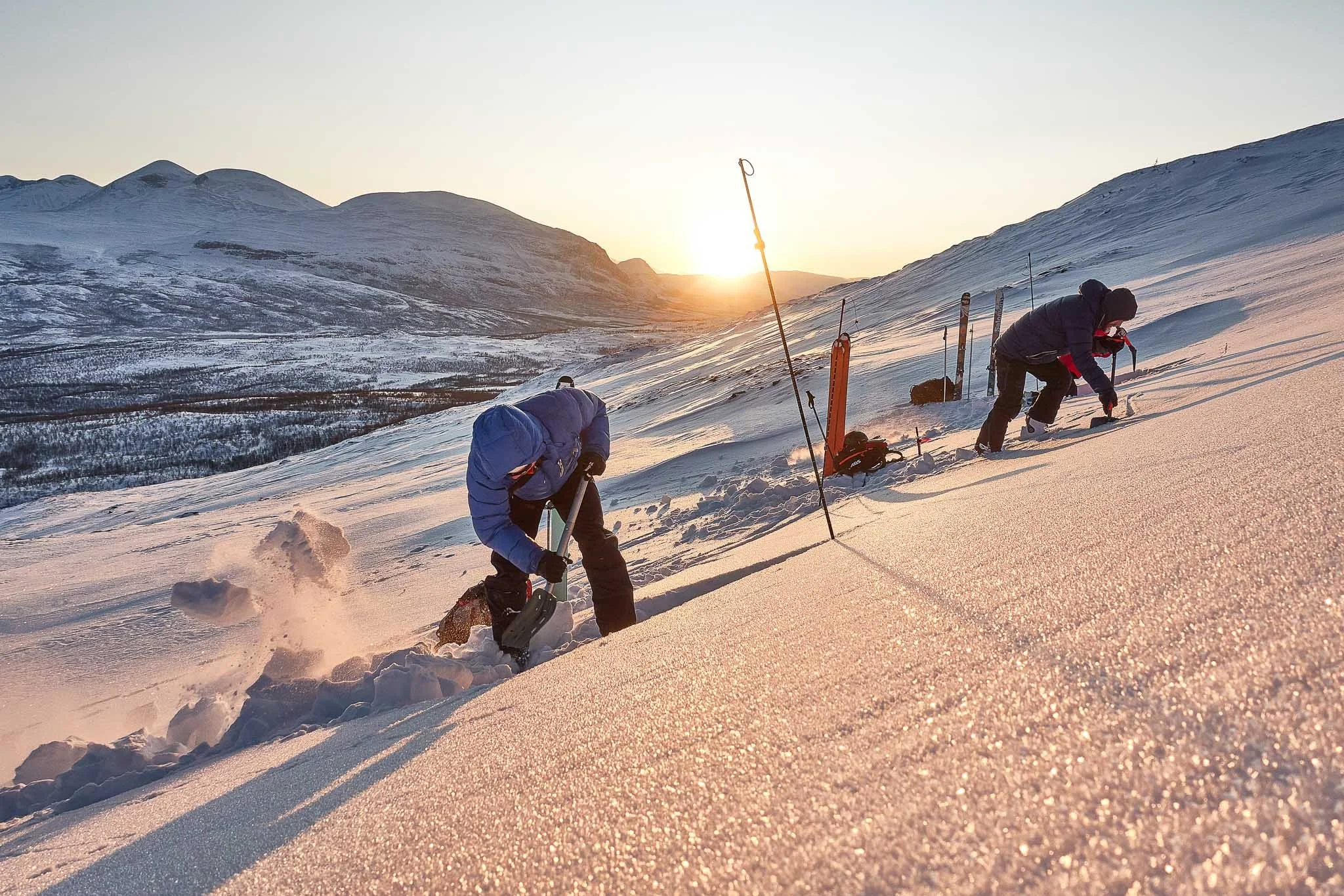 To personer som bruker snøspader på en snødekt fjellskråning under solnedgang.