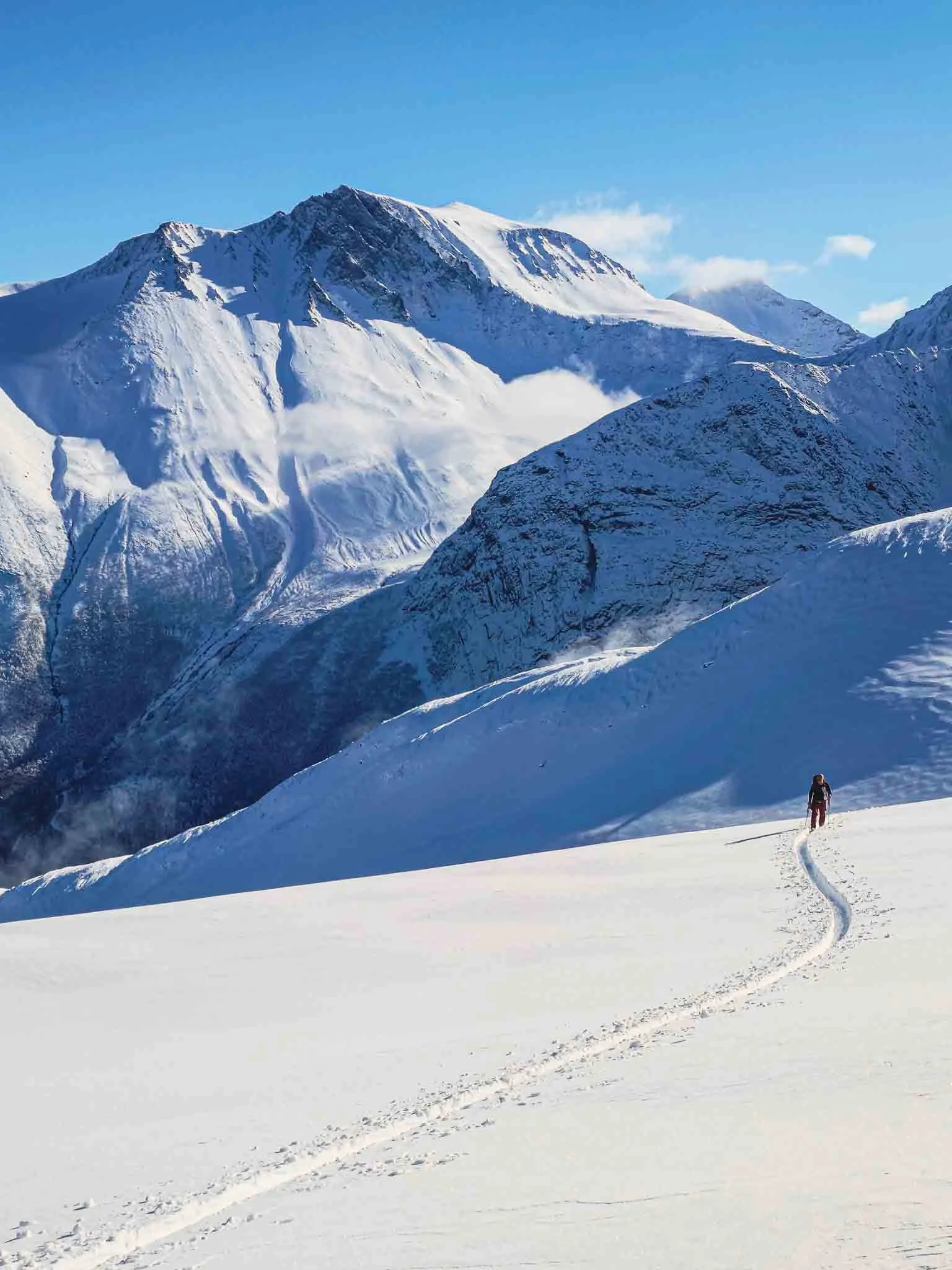 En skidåkare vandrar i snön med höga, snöklädda berg i bakgrunden under en klarblå himmel.