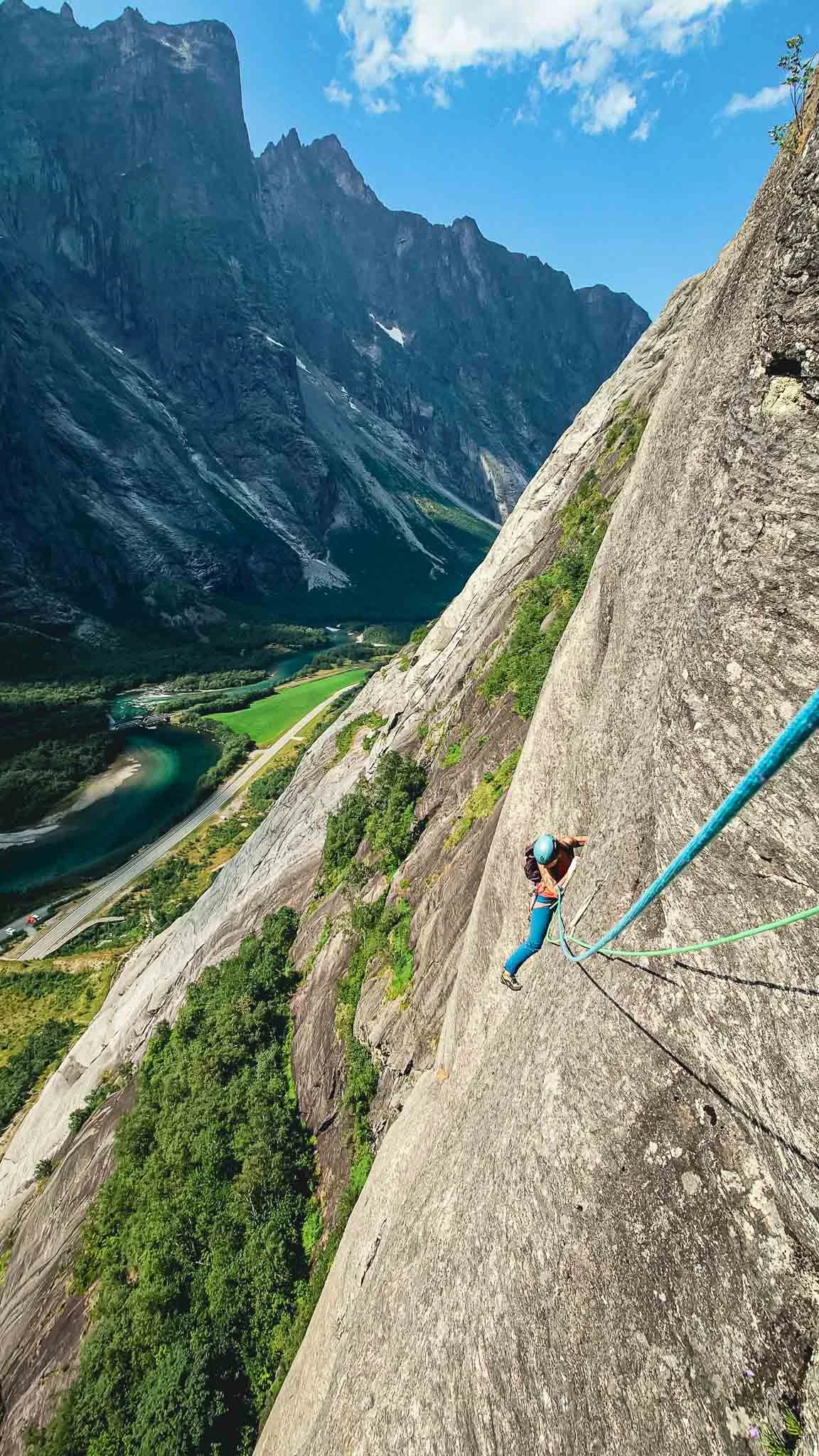 En person klatrer på en bratt fjellvegg med utsikt over en elv og grønne åser i en fjellfjord.