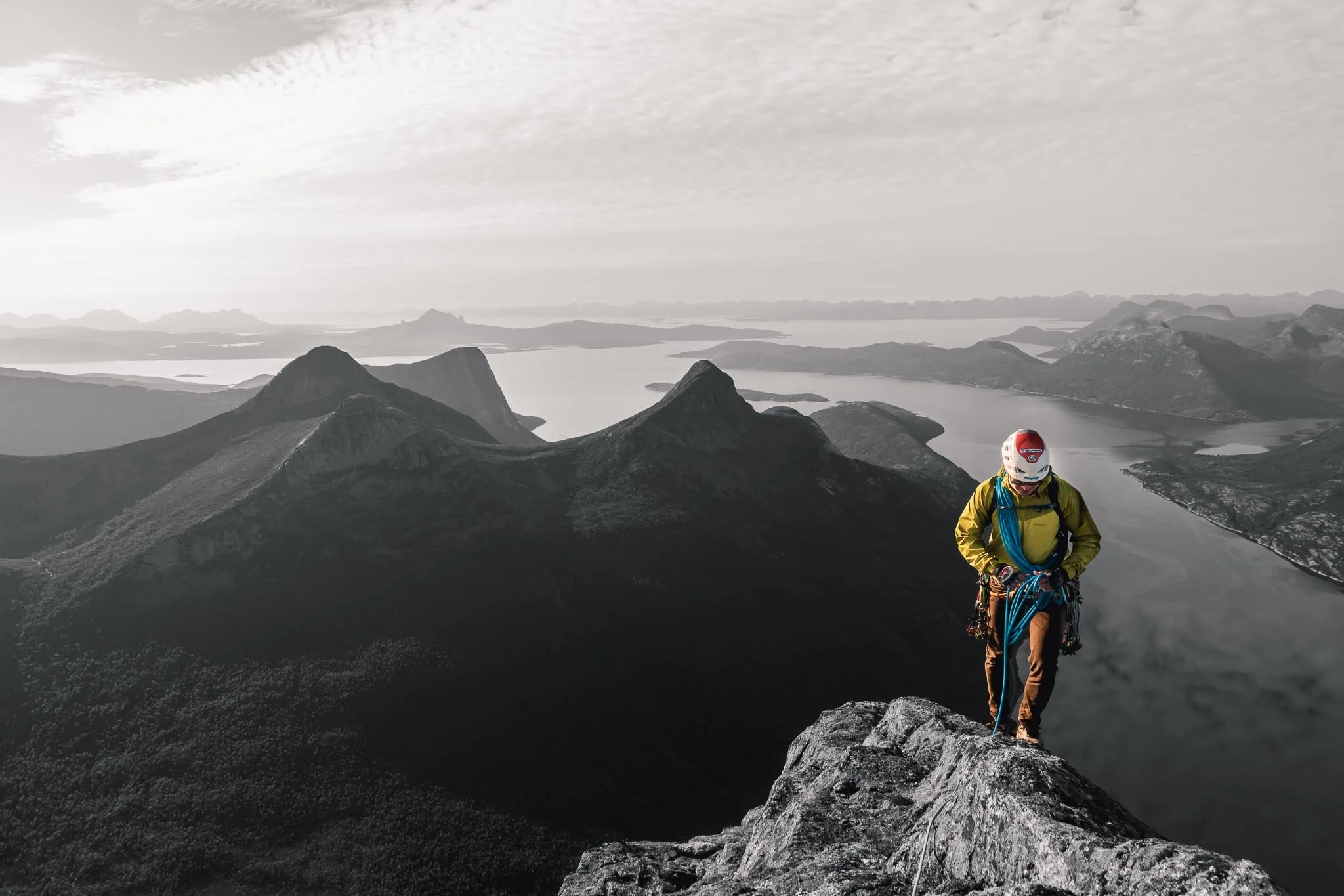 En bergsklättrare står på en bergstopp med berg och sjöar i bakgrunden, delvis i svartvitt, med klätterutrustning och hjälm.