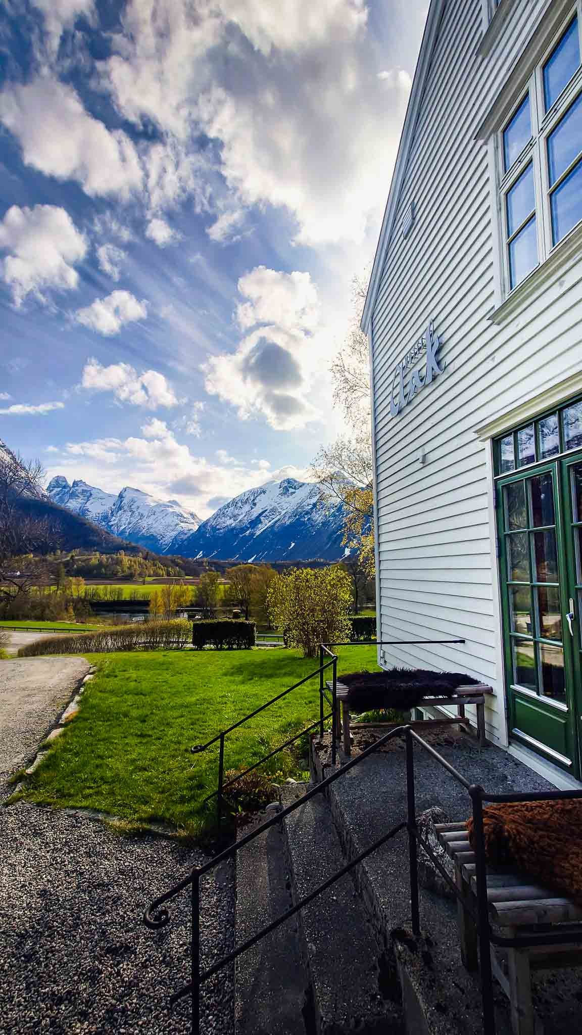 Et utsikt over fjell og landskap med snødekte topper, grønne marker, en hvit bygning med grønne vinduer og dør, og en liten terrasse med benker i Norge.
