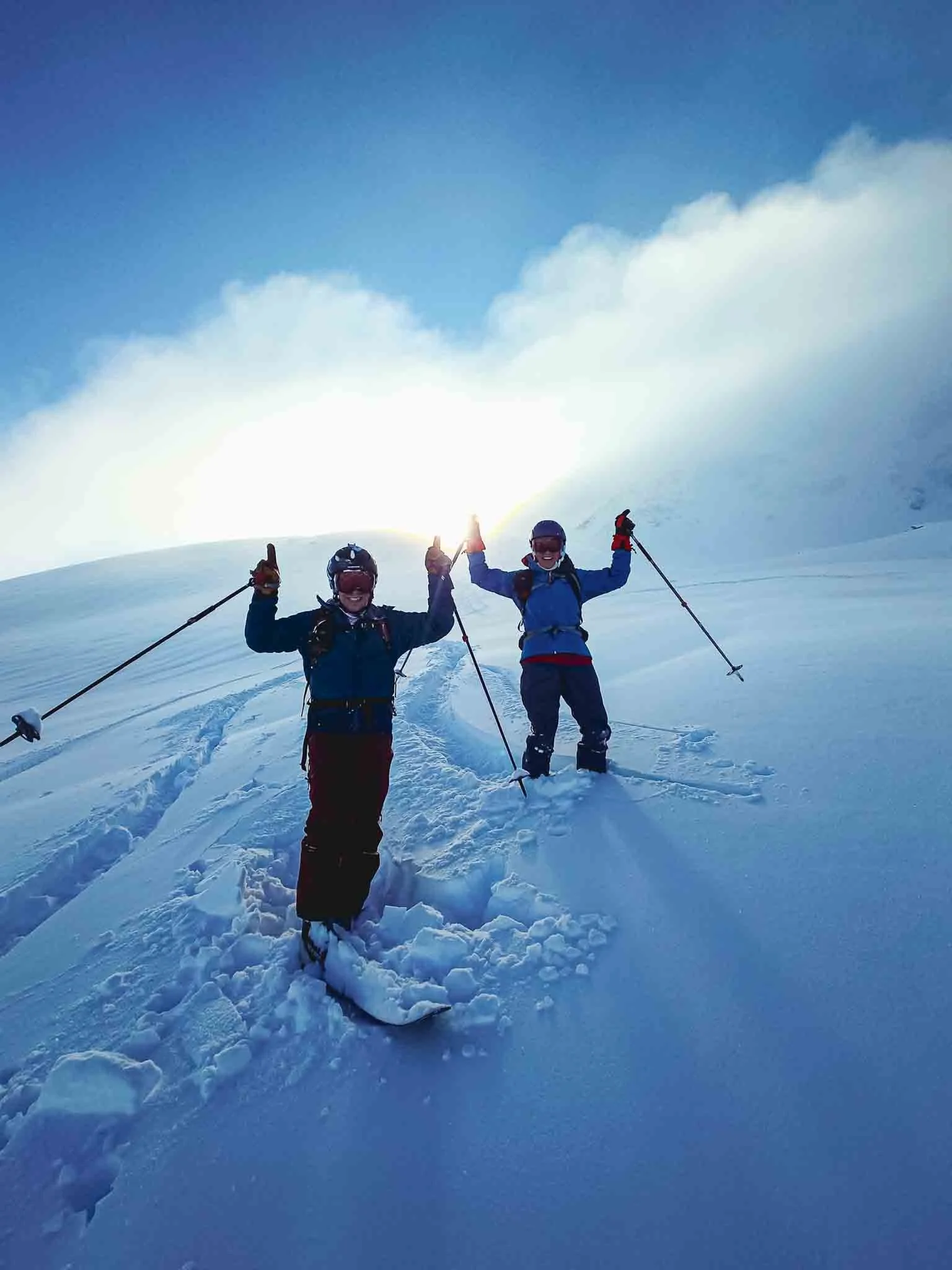 Två personer på en snötäckt bergstopp, en med skidor och en med kikare, firar med upphöjda händer under en blå himmel och solnedgång.