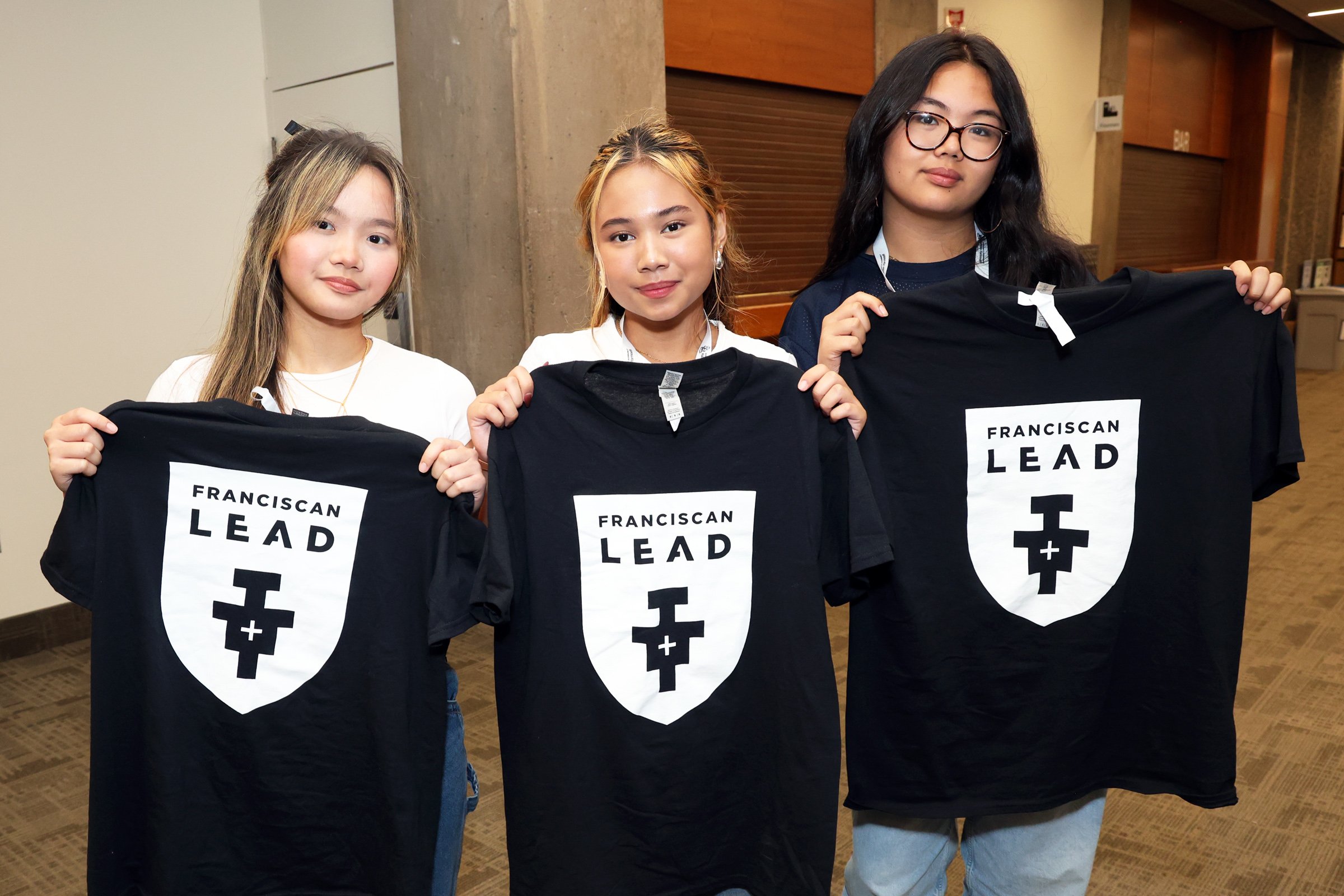 Three young women standing indoors, each holding a black T-shirt with a white shield logo. The shirts have the words "Franciscan Lead" and a shield emblem.