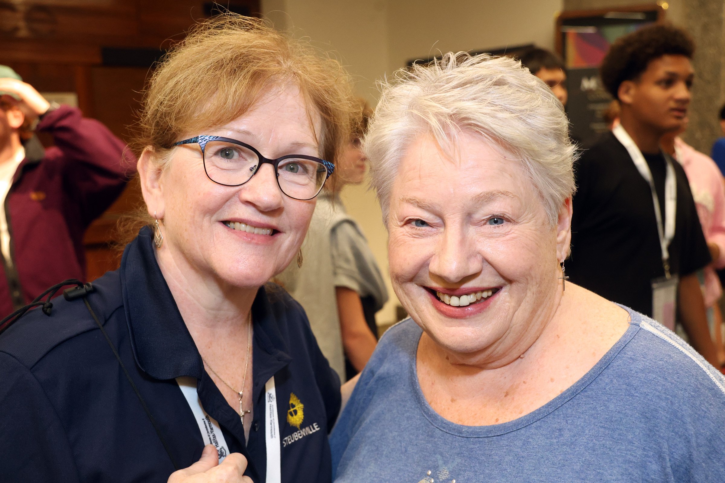Two smiling women, one with glasses and red hair and the other with short gray hair, posing for the camera at an indoor event with a crowd in the background.