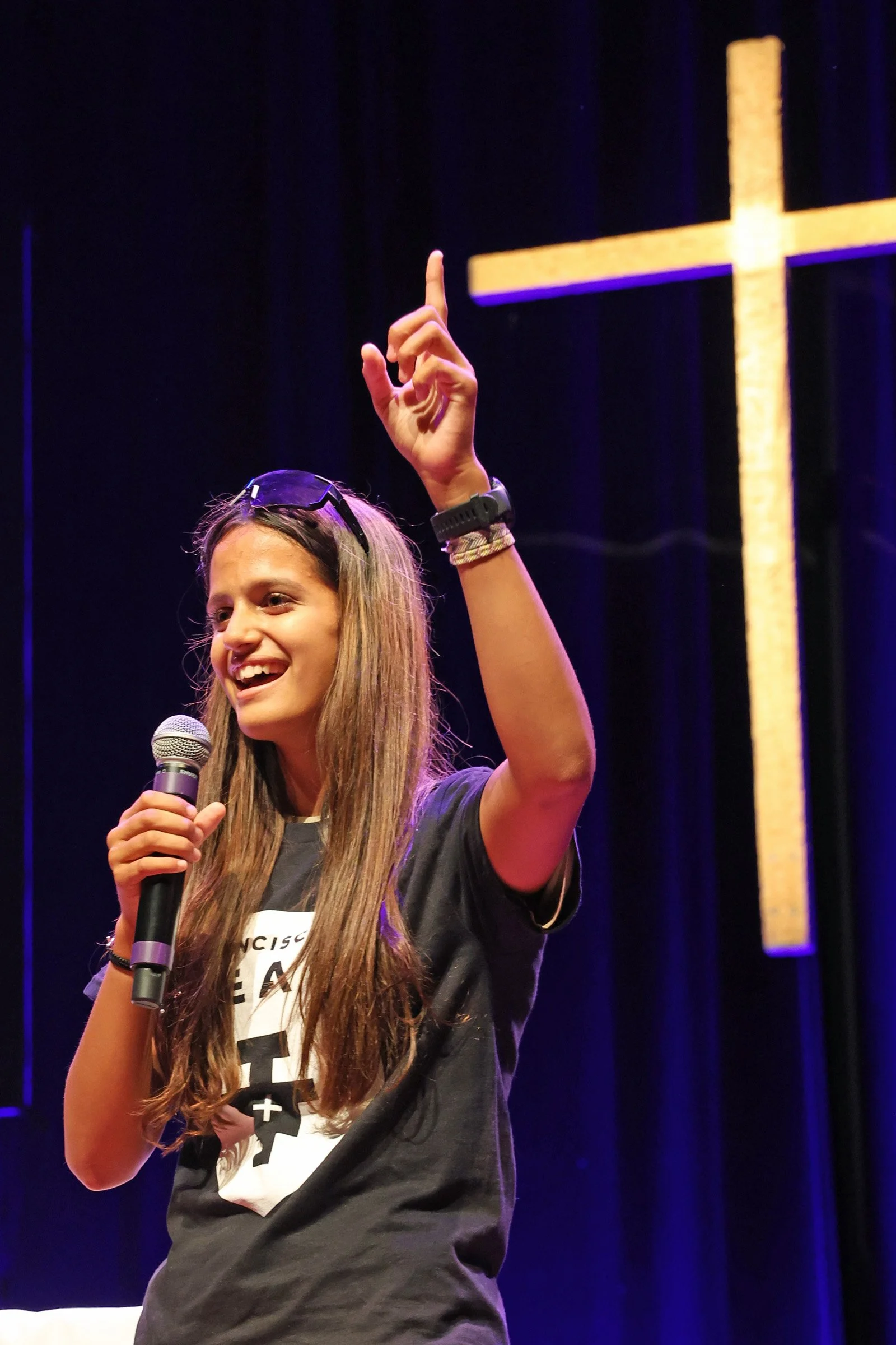 A young woman with long hair holding a microphone and speaking on stage, with a cross illuminated in the background.