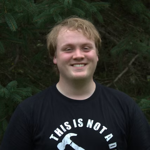 Young man with blonde hair smiling outdoors with green trees in the background.