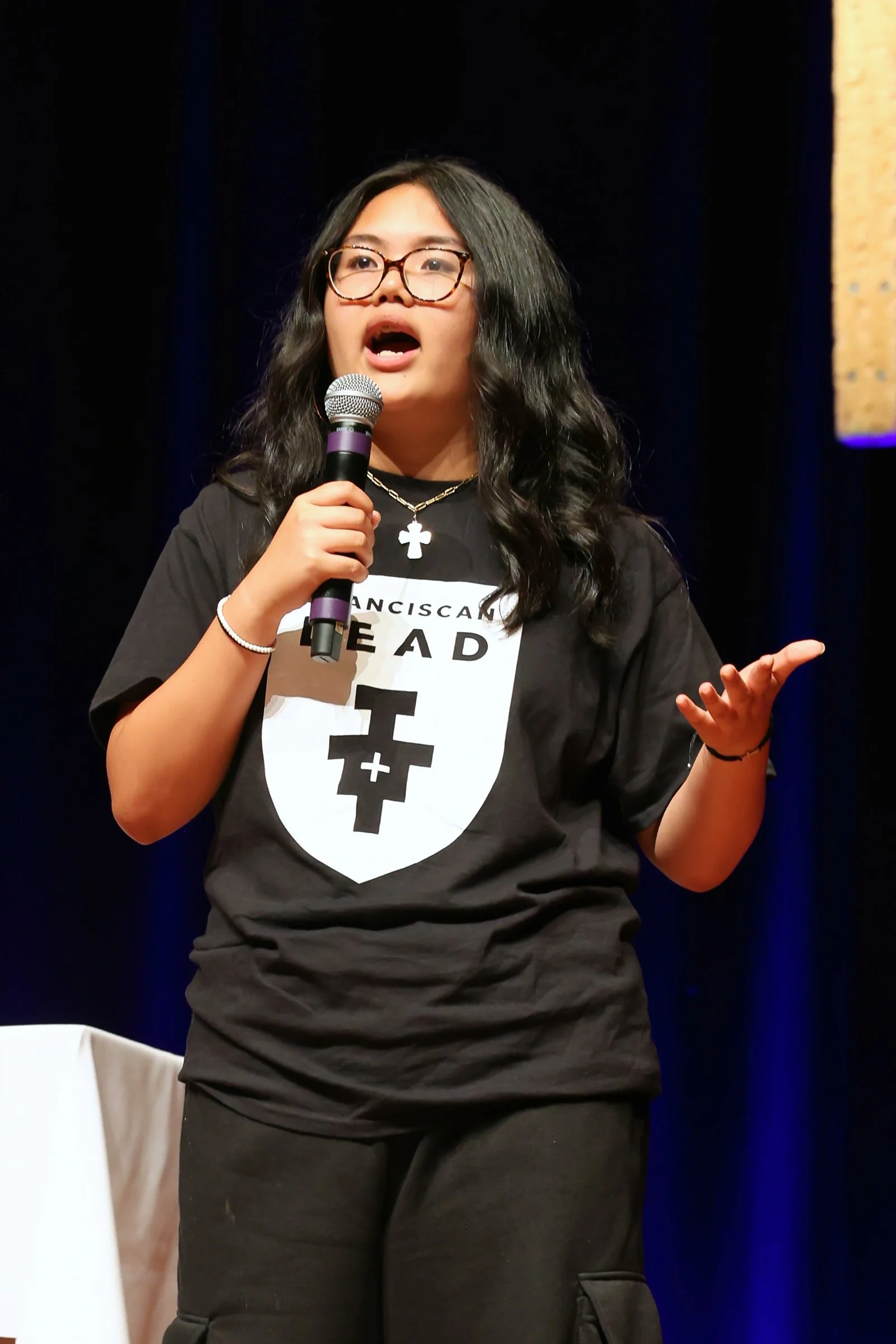 Young woman speaking into a microphone on stage, wearing glasses, a black t-shirt with a logo, and a cross necklace.