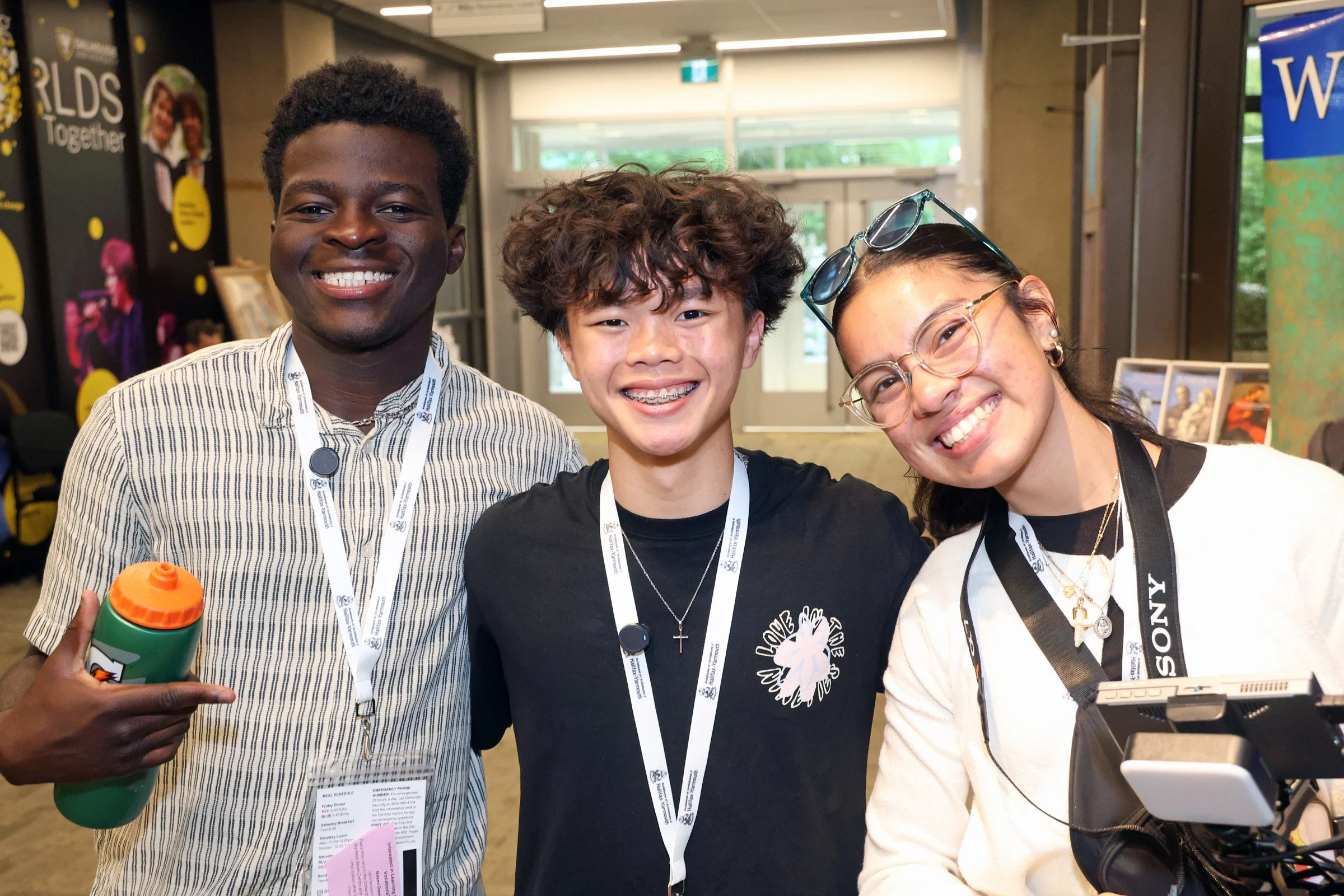 Three young people smiling and posing for a photo at an indoor event. The person on the left is holding a green water bottle with an orange lid, the person on the right has sunglasses on her head and a camera in front of her. All three are wearing lanyards with badges.