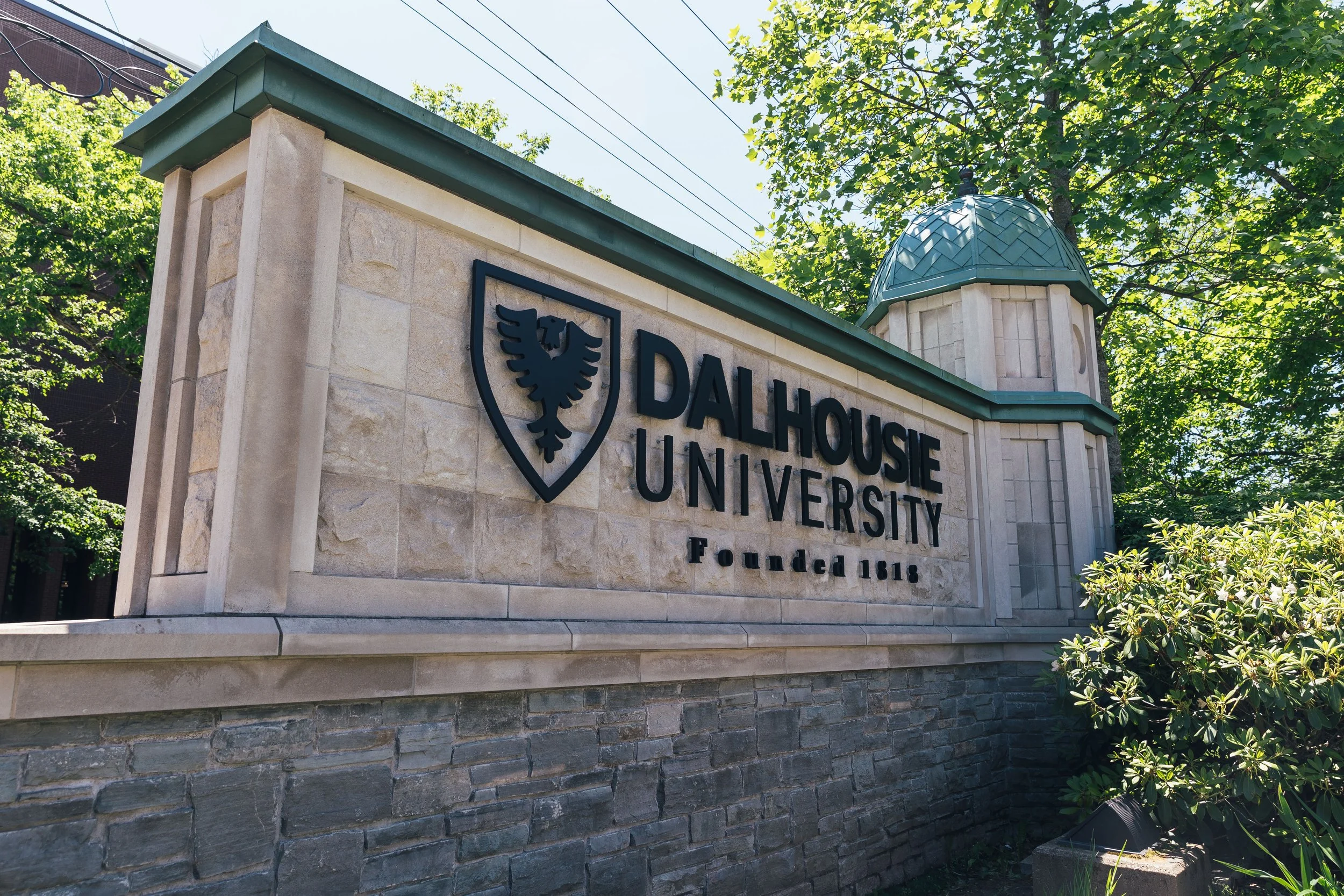 The entrance sign of Dalhousie University, featuring a shield with a black eagle, the university’s name, and its founding year 1818, surrounded by greenery and a blue sky.