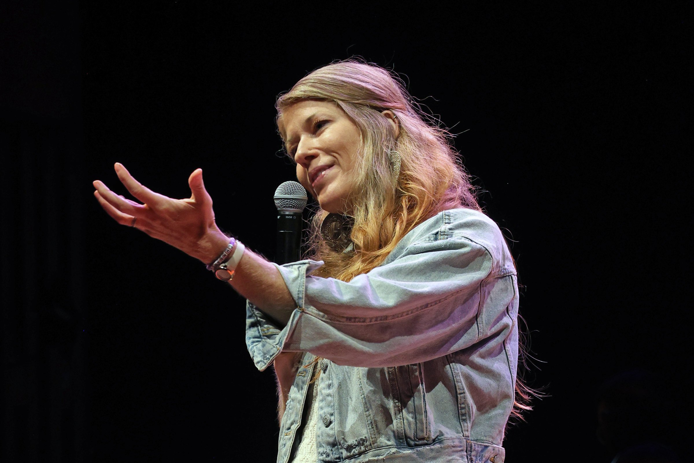 A woman with long, wavy blonde hair holding a microphone and speaking on stage against a black background.