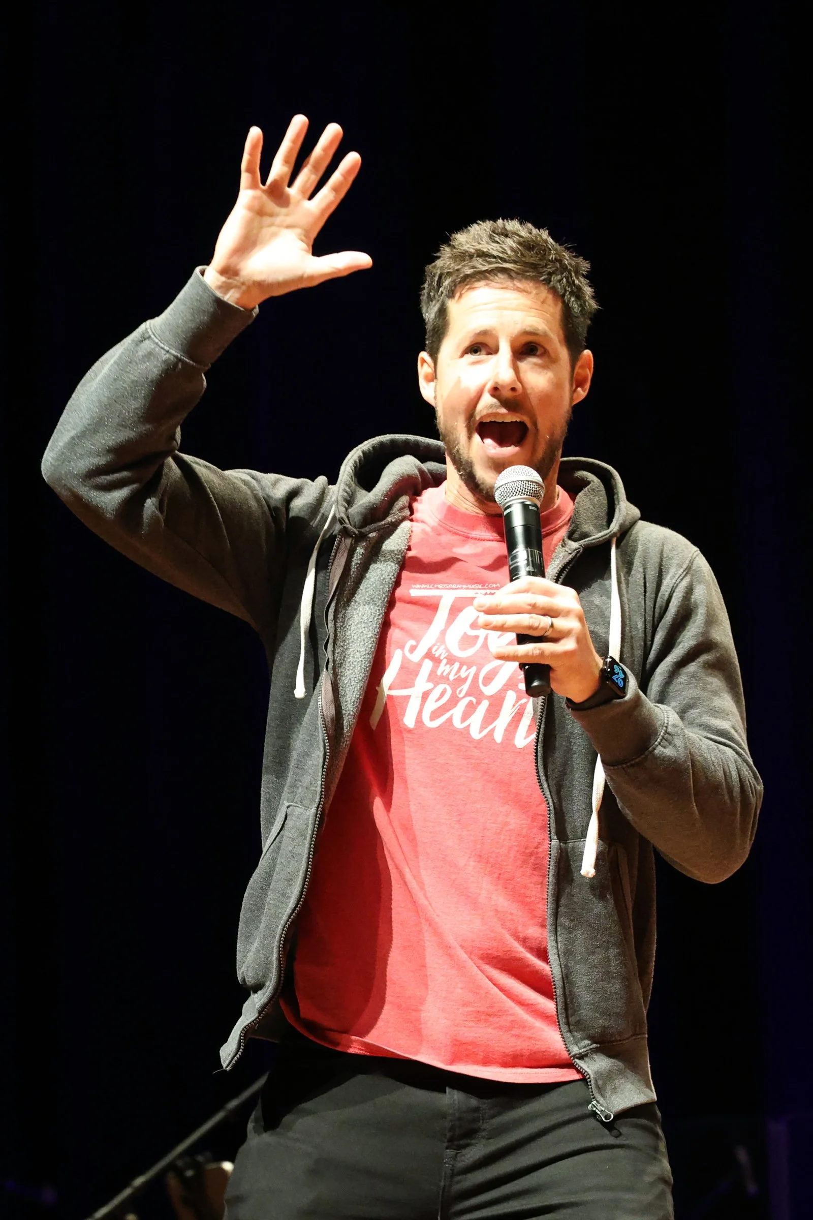 Man speaking into a microphone on stage, wearing a red t-shirt and gray hoodie, with his right hand raised.