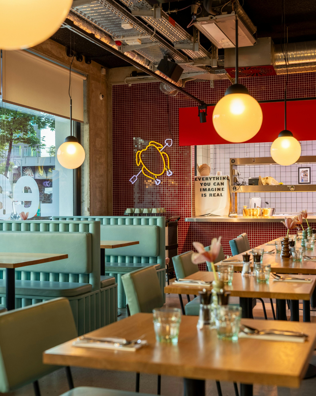 Interior of a restaurant with modern decor, including pastel green booths, wooden tables with glass and cutlery, and a red tiled wall with neon art. Large round pendant lights hang from the ceiling, and there's a window showing trees outside. In the background, a sign reads, 'Everything you can imagine is real.'