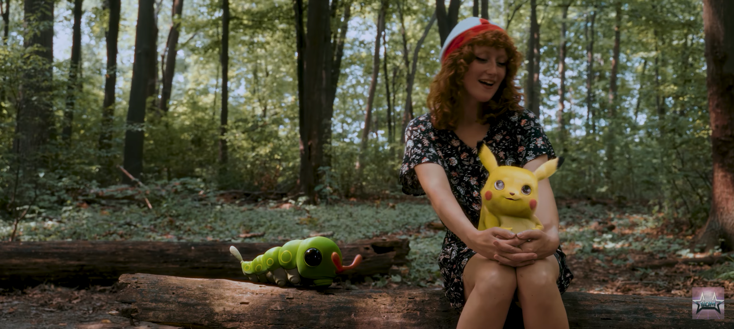 A woman in a floral dress sitting on a log in a forest, holding a Pikachu, with a large caterpie on the log beside her.