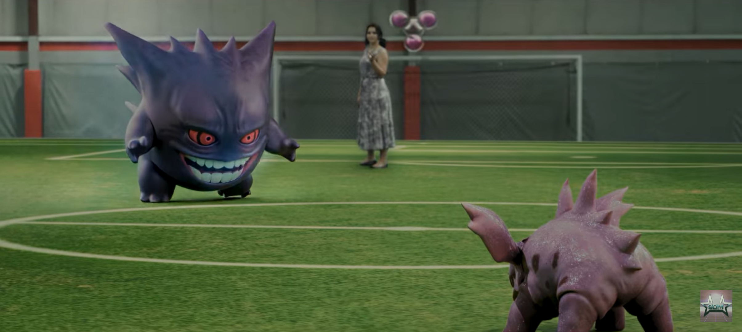 A scene from a Pokémon movie showing two Pokémon, Gengar and Sandslash, on an indoor sports field with green artificial turf. There is a woman in the background near a cluster of pink and white balloons, standing near a goalpost.