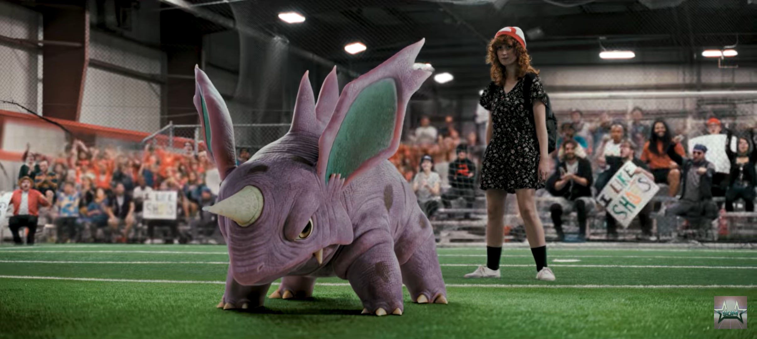 A woman with red curly hair wearing a black floral dress and a Santa hat stands on an indoor sports field, facing a large, purple, cartoonish rhinoceros with exaggerated features and vibrant green ears, surrounded by a crowd of spectators holding sig