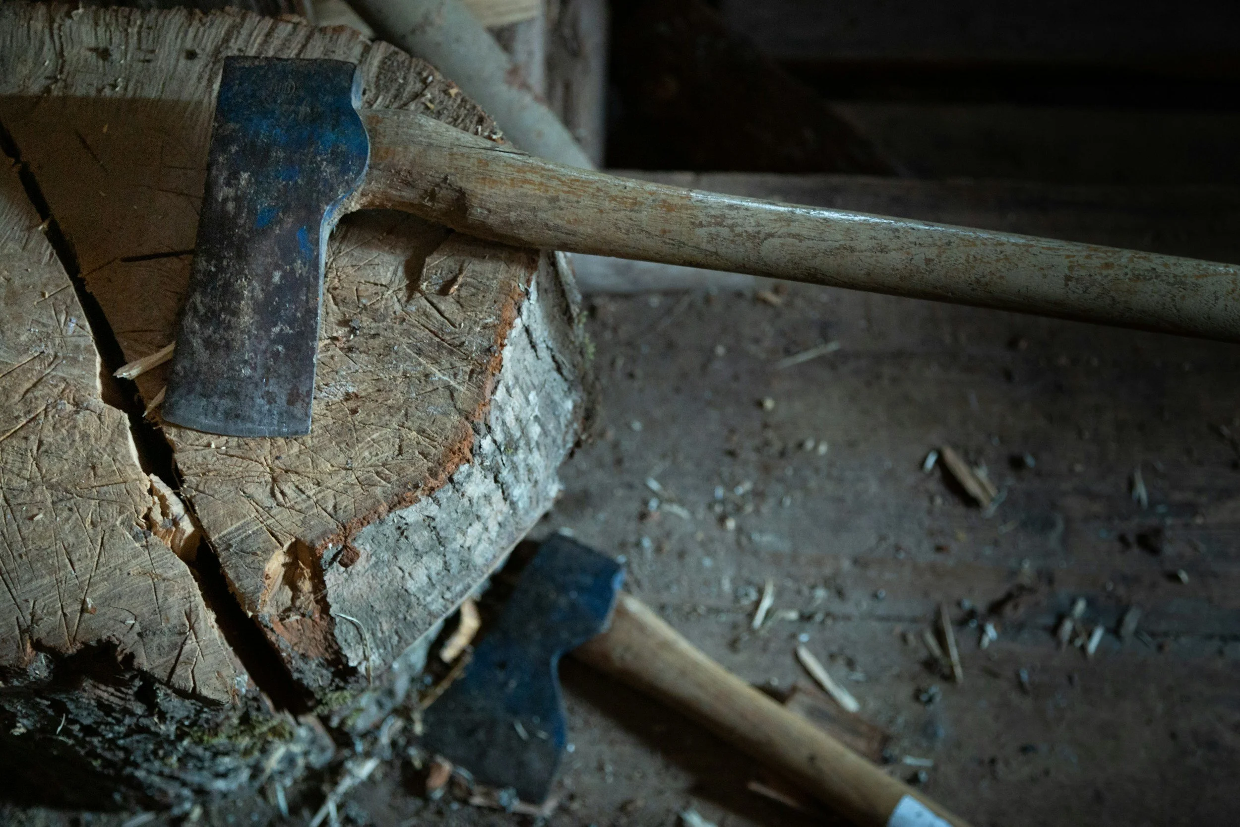 An axe with a worn handle and a rusty blade resting on a chopped piece of wood, with another axe lying nearby on a dirt floor.