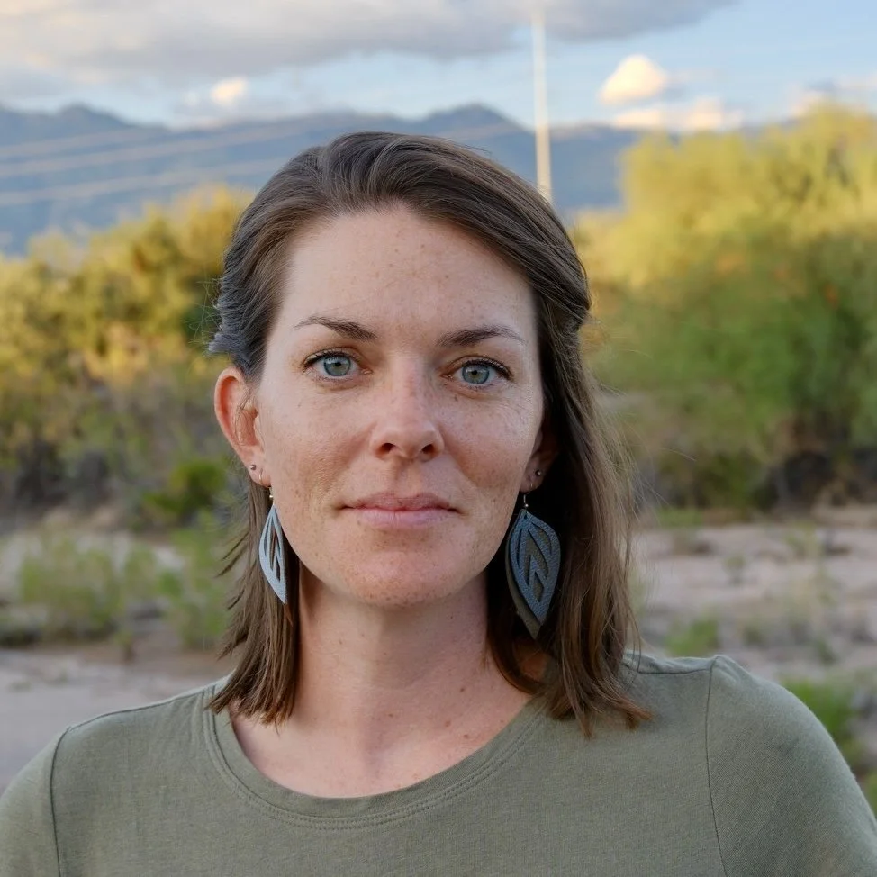 A woman with shoulder-length brown hair, blue eyes, wearing large leaf-shaped earrings and a green shirt, outdoors with trees and mountains in the background.