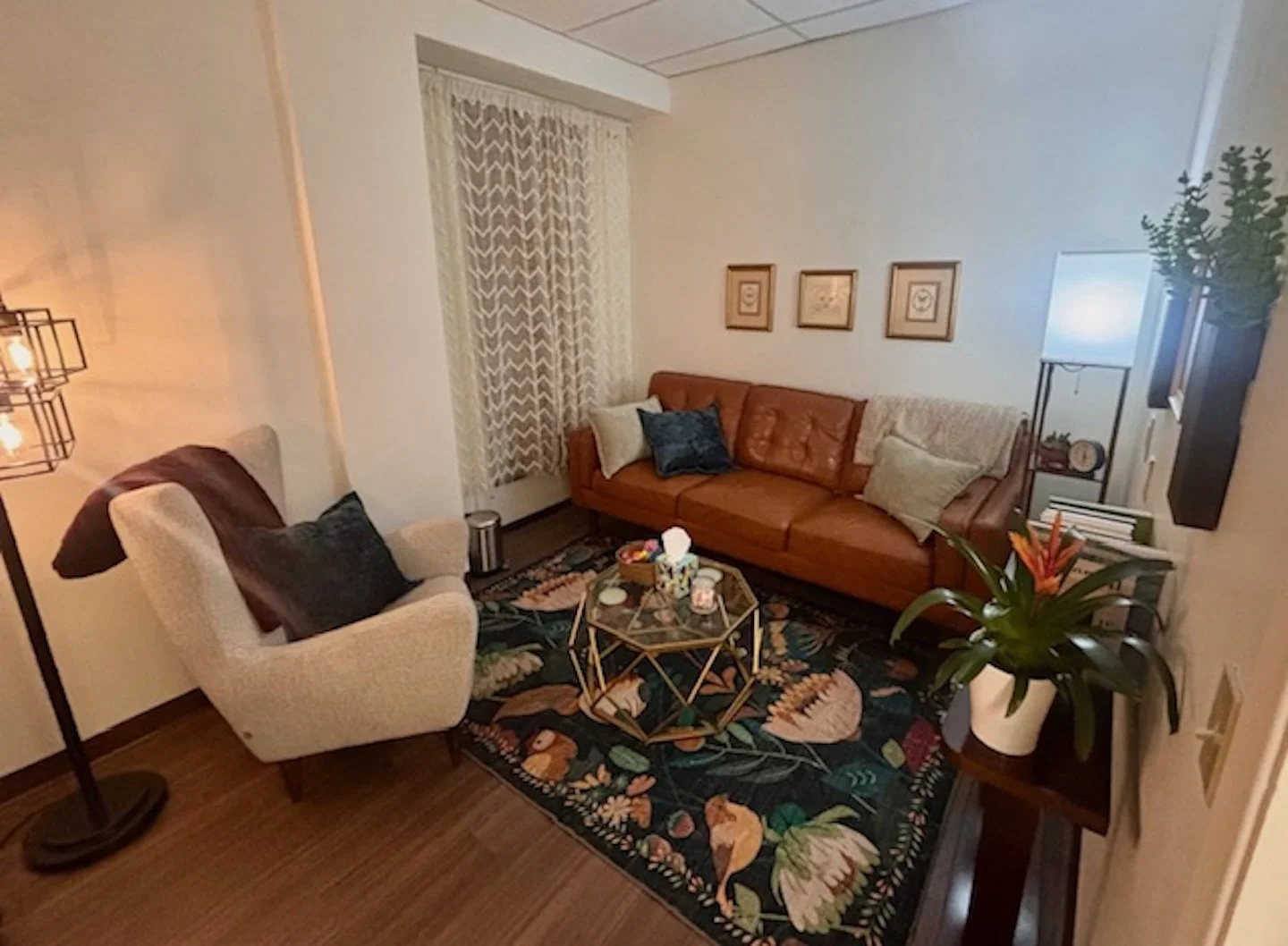 Cozy living room with a tan armchair, a brown leather couch, a black framed floor lamp, paintings on the wall, a floral rug, a glass coffee table with decorative items, and a houseplant with orange flowers.