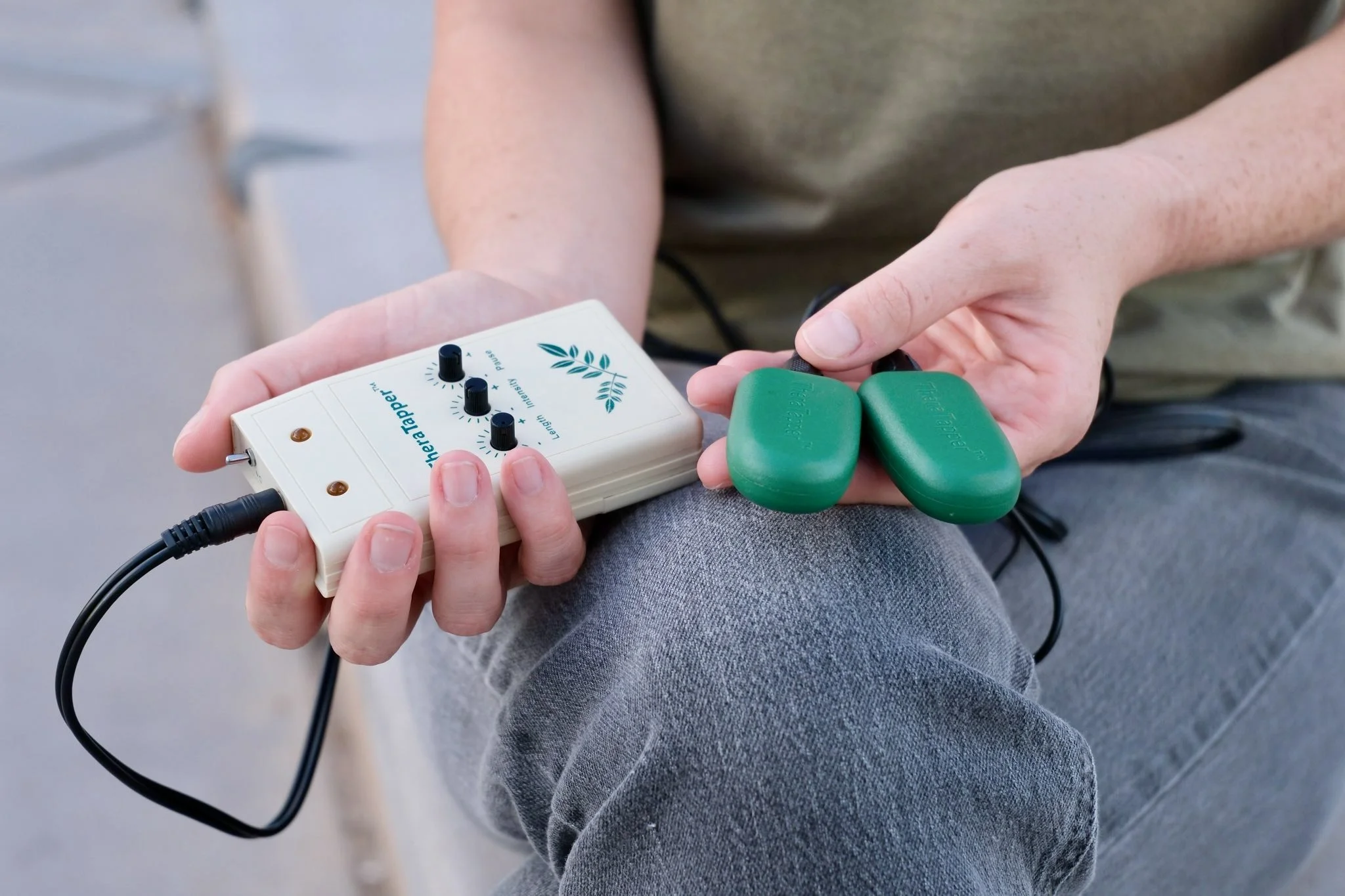 Person holding EMDR tappers, which are a white electronic device with black knobs and two green footpads connected by a wire.