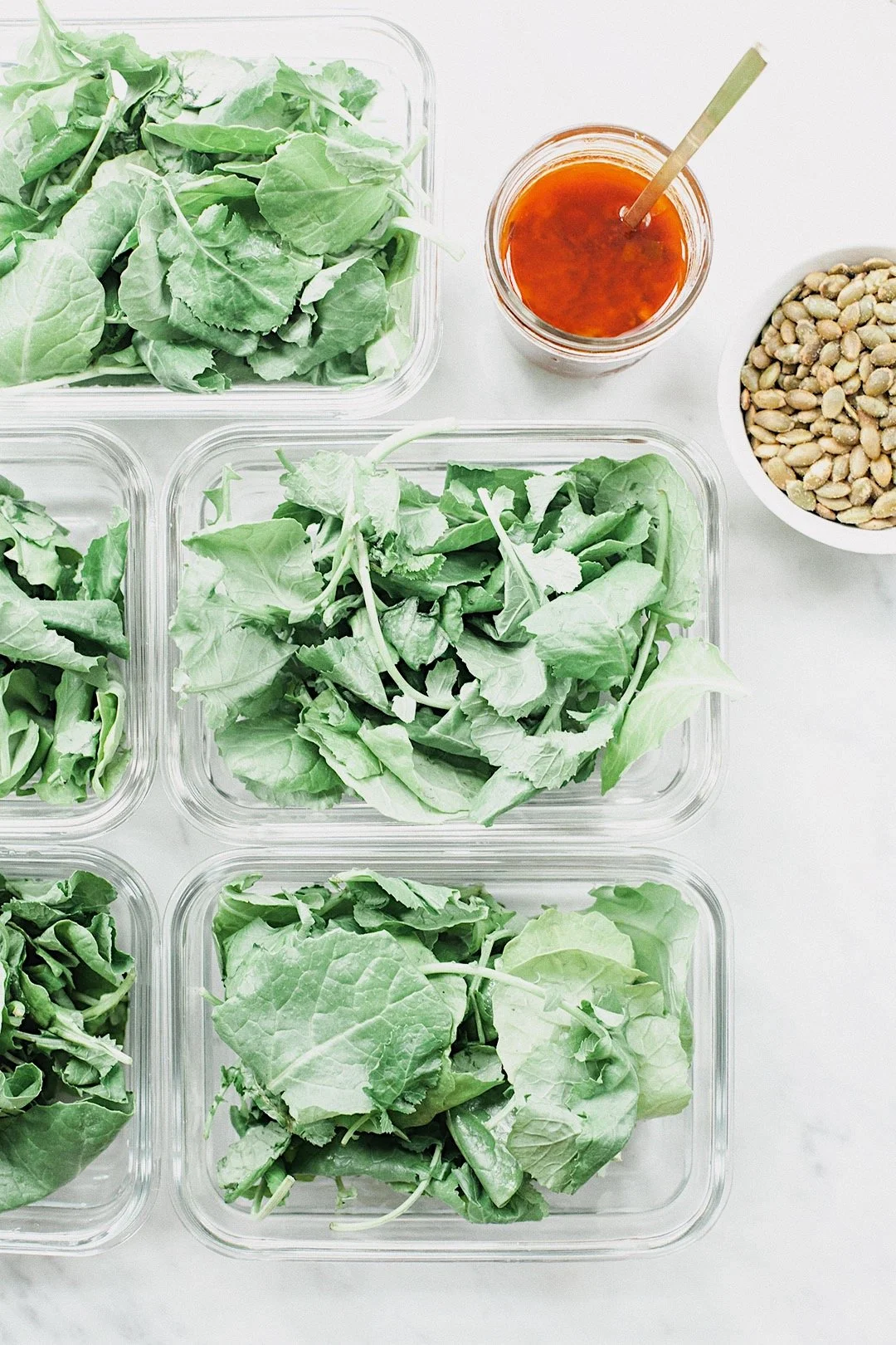 Salad greens portioned into meal prep containers with dressing and pumpkin seeds.