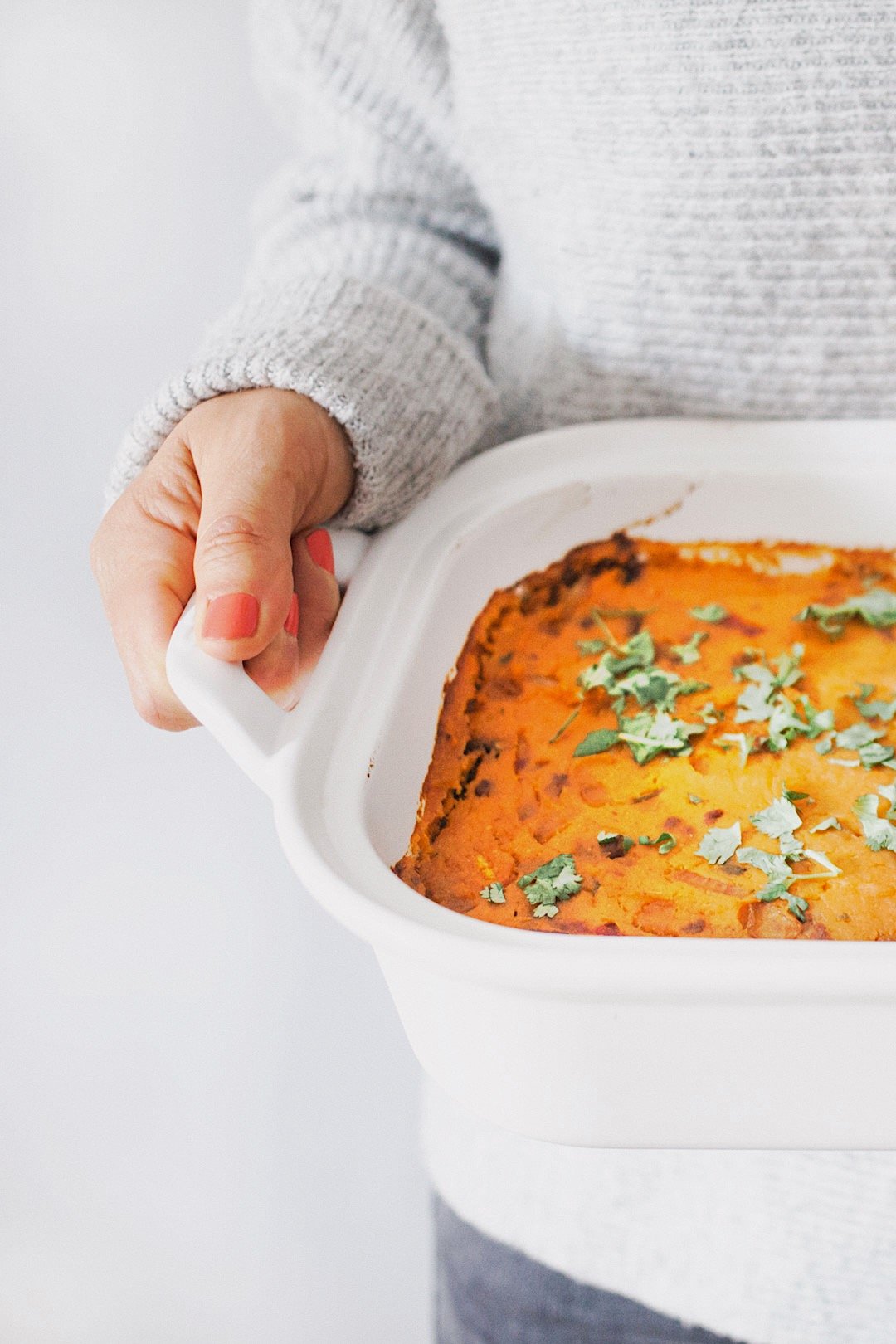 Person holding a white ceramic baking dish with a baked casserole garnished with fresh herbs.