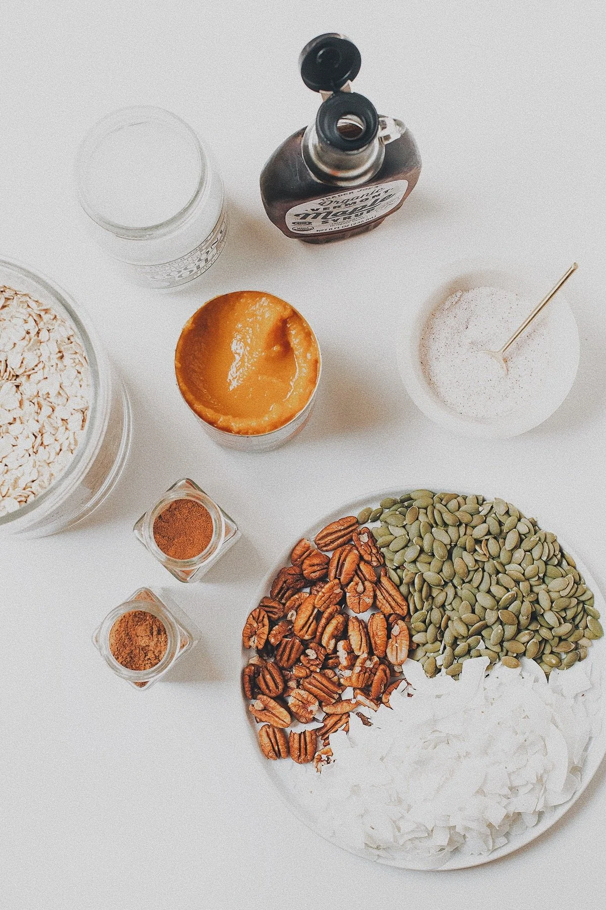Ingredients for pumpkin granola including oats, pumpkin purée, pecans, pumpkin seeds, coconut, maple syrup, and spices.