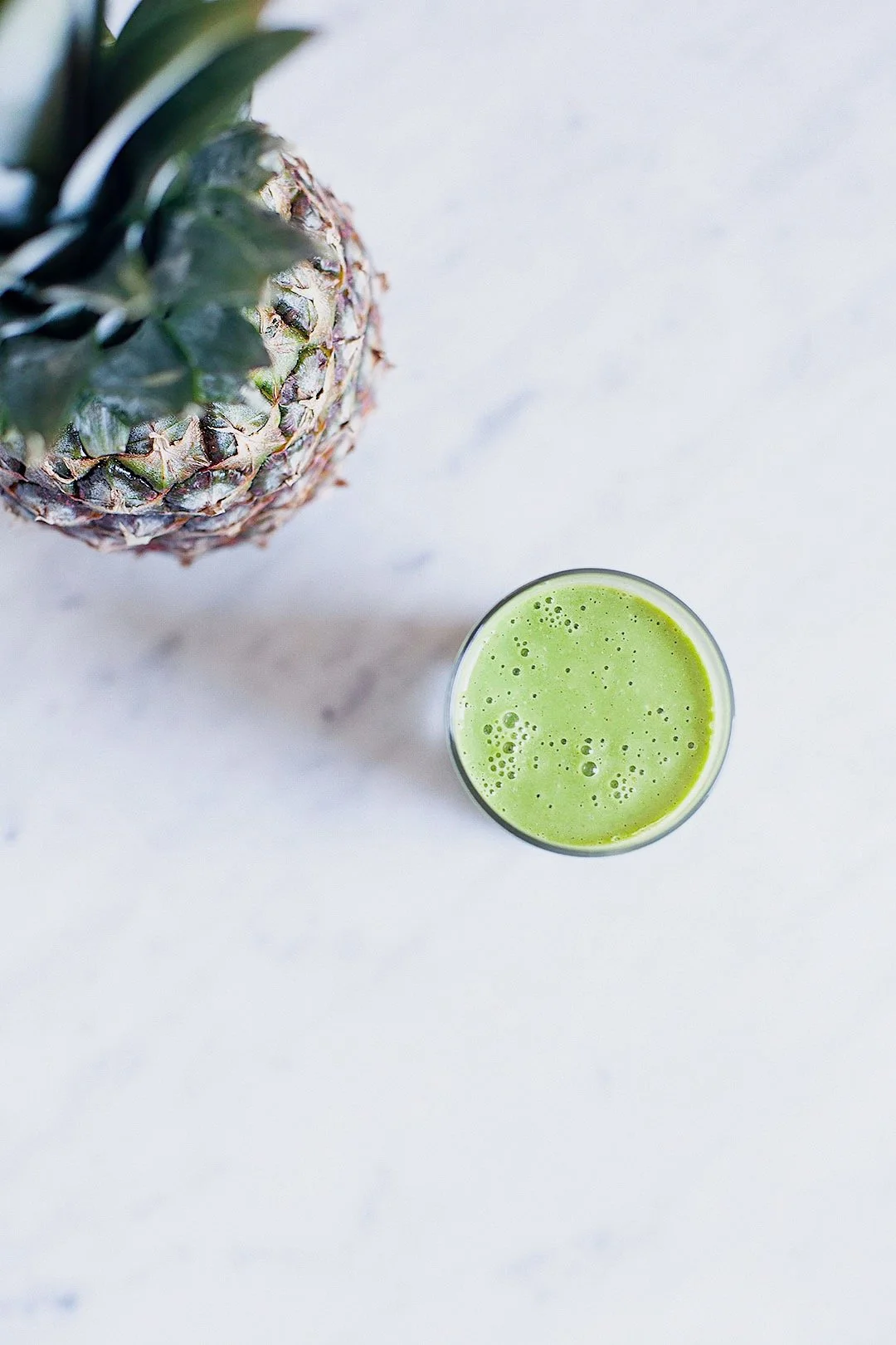 Vibrant green pineapple and kale smoothie in a glass on a white marble surface.