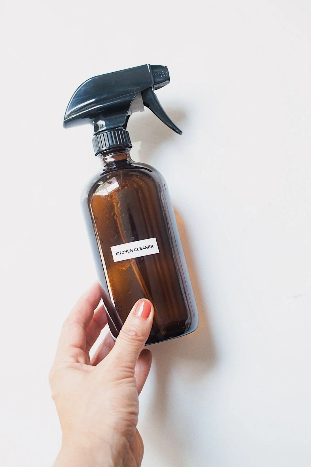 Hand holding a glass spray bottle of non-toxic kitchen cleaner on a white countertop.