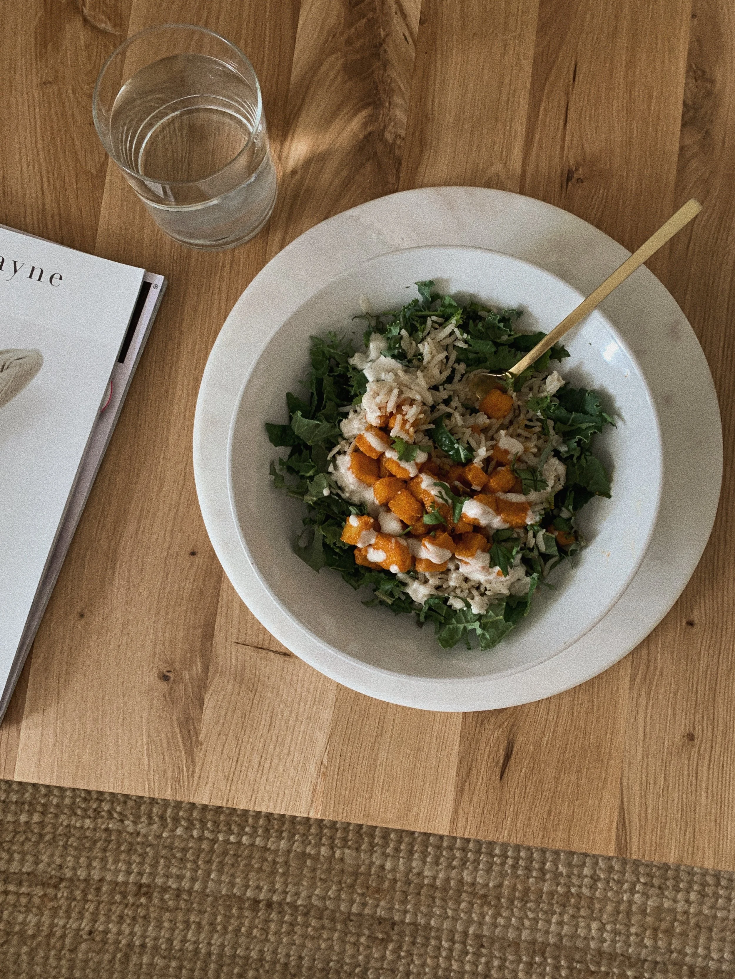 A white bowl of salad on a wooden table, with a fork, beside a glass of water and a book.