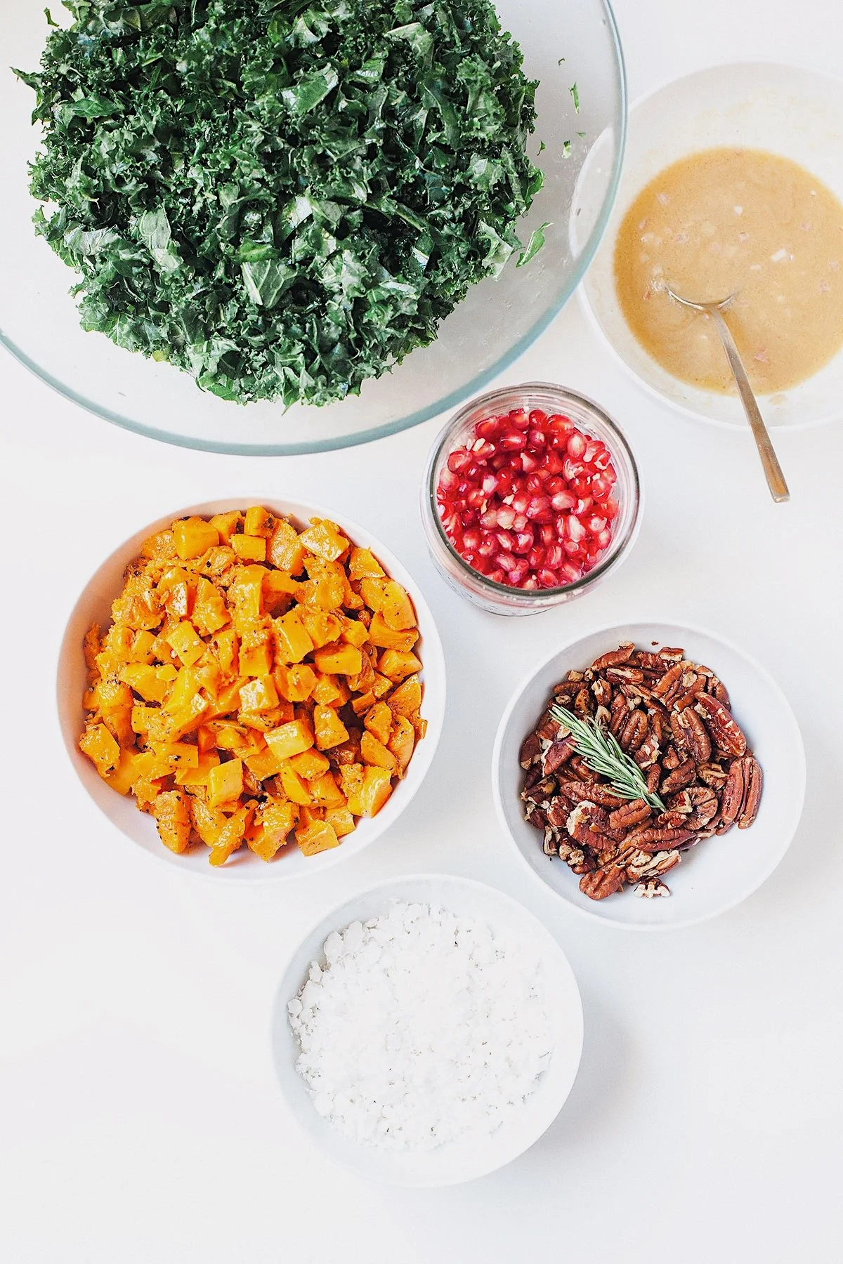 Ingredients for Thanksgiving kale salad on a white surface: kale, squash, pomegranate, dressing, and pecans.