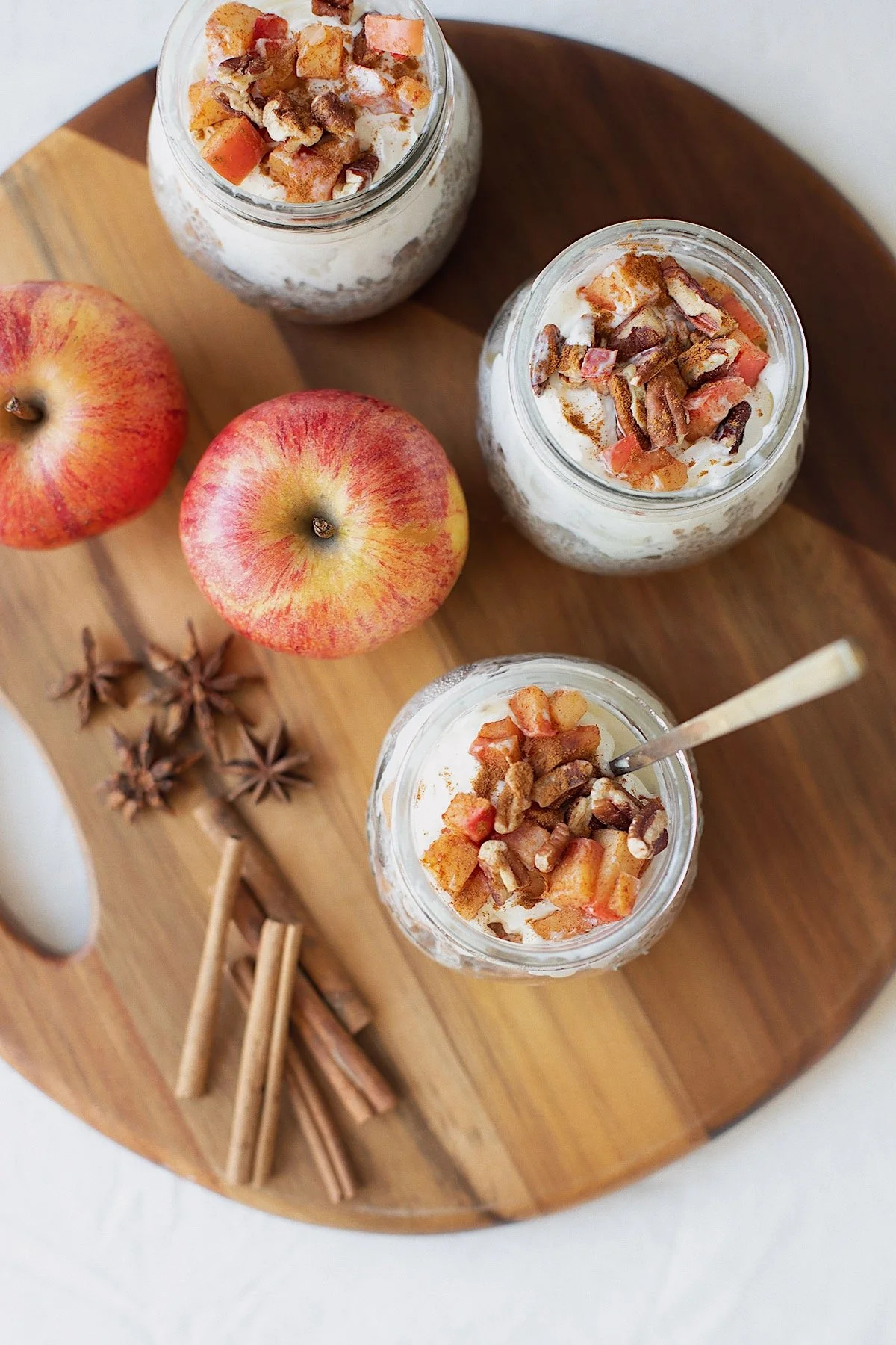 Overhead view of apple pie chia pudding jars topped with sautéed apples and pecans on wooden board with fresh apples and cinnamon sticks.