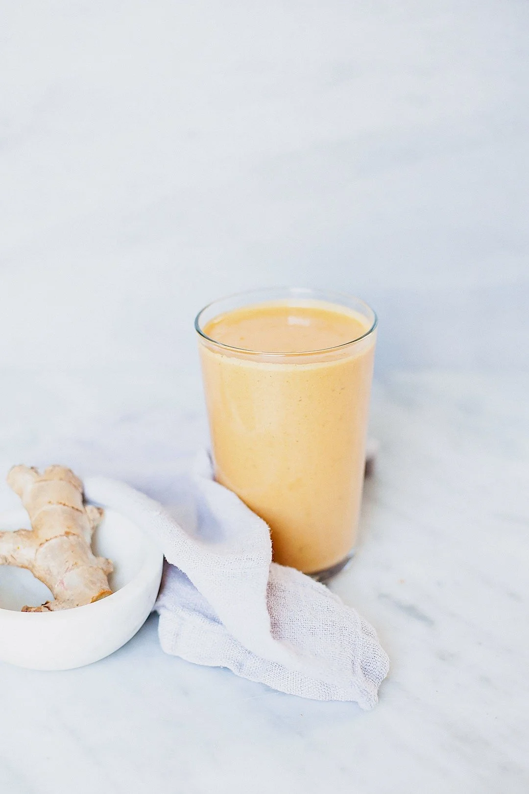 Bright yellow mango and turmeric smoothie in a glass with fresh ginger on a white marble surface.