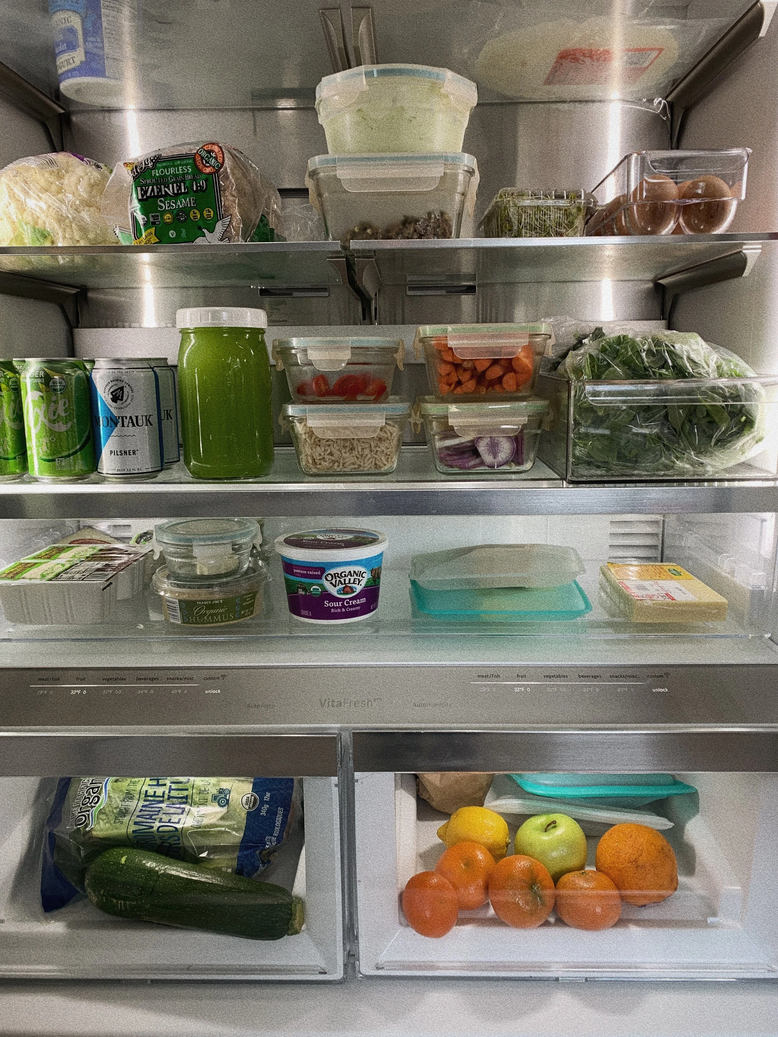 Inside of a refrigerator showing various food items, including oranges, apples, lemons, cucumbers, cherry tomatoes, leafy greens, eggs, hummus, organic valley sour cream, canned beverages, and plastic containers with vegetables and other ingredients.