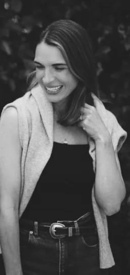 Black and white photo of a woman smiling, wearing a sleeveless top and a cardigan, standing outdoors with trees in the background.
