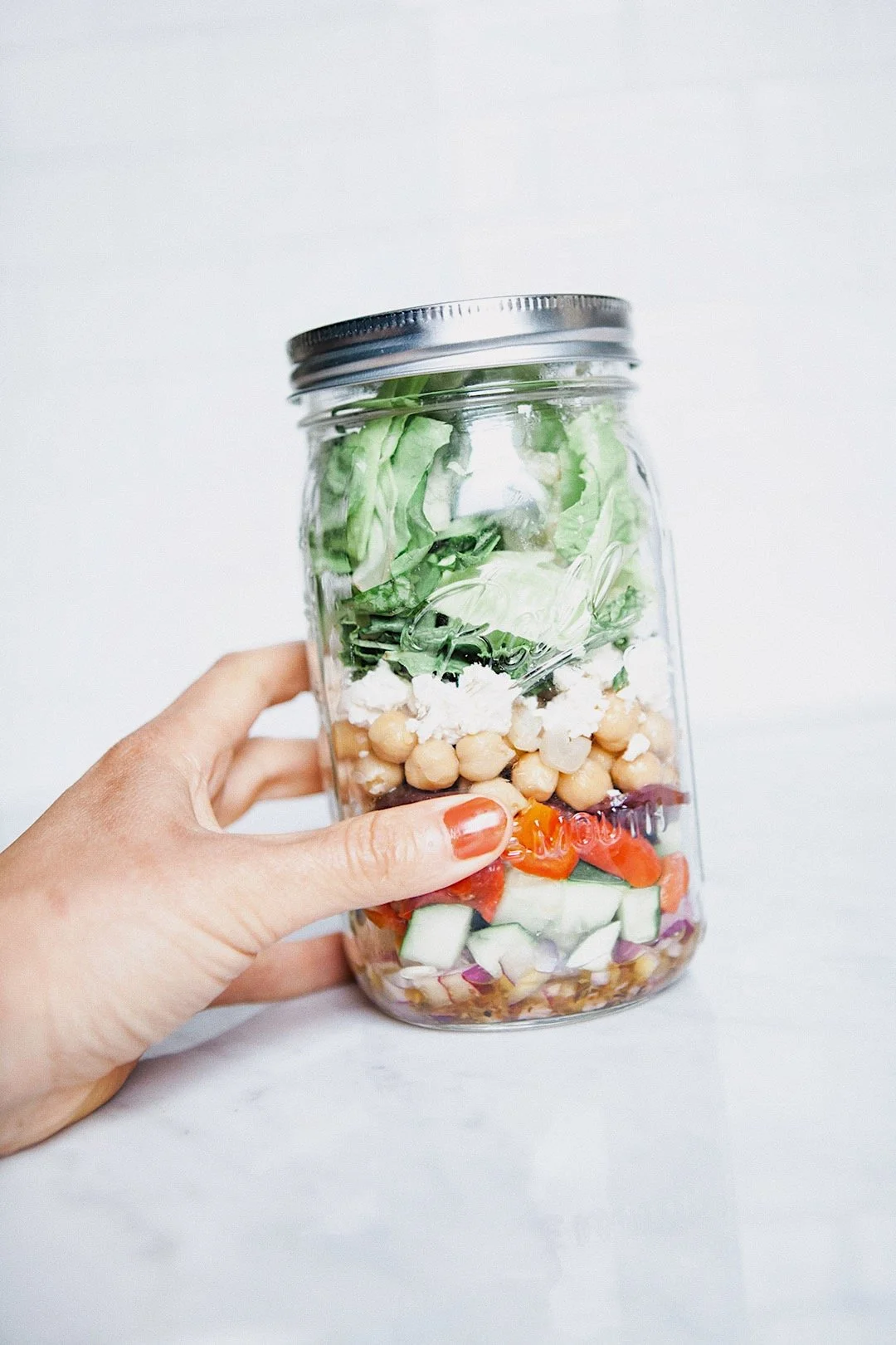 Mason jar salad layered with dressing, vegetables, chickpeas, feta, and greens.
