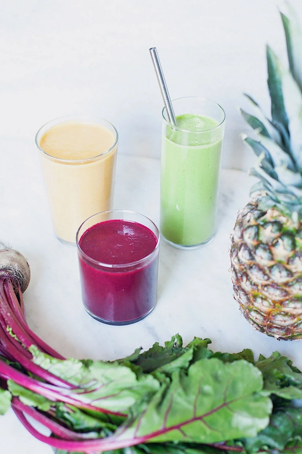 Three colorful smoothies for healthy digestion on a white surface with pineapple, kale, and beets.