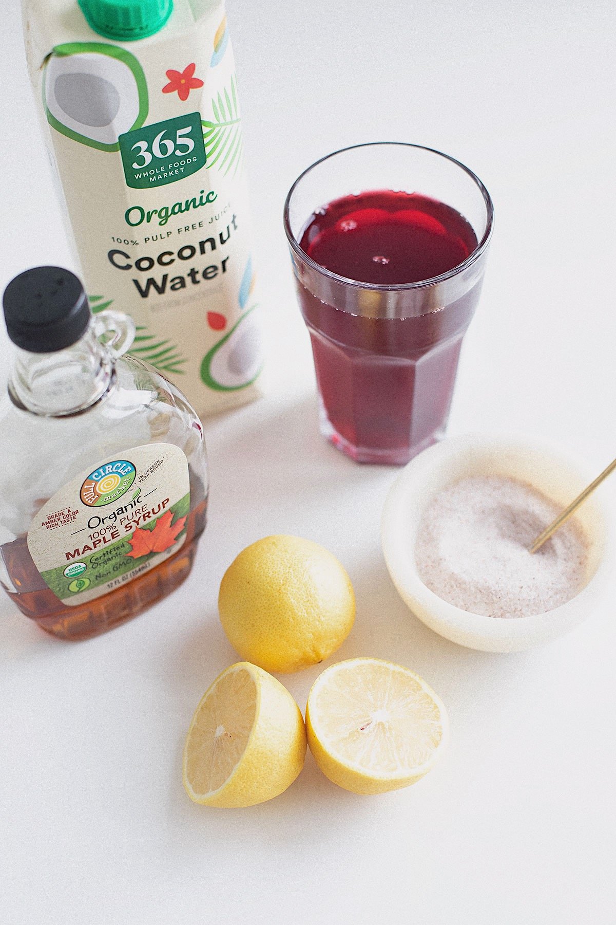 Ingredients for a natural electrolyte drink: coconut water, lemon, cranberry juice, maple syrup, and mineral salt.