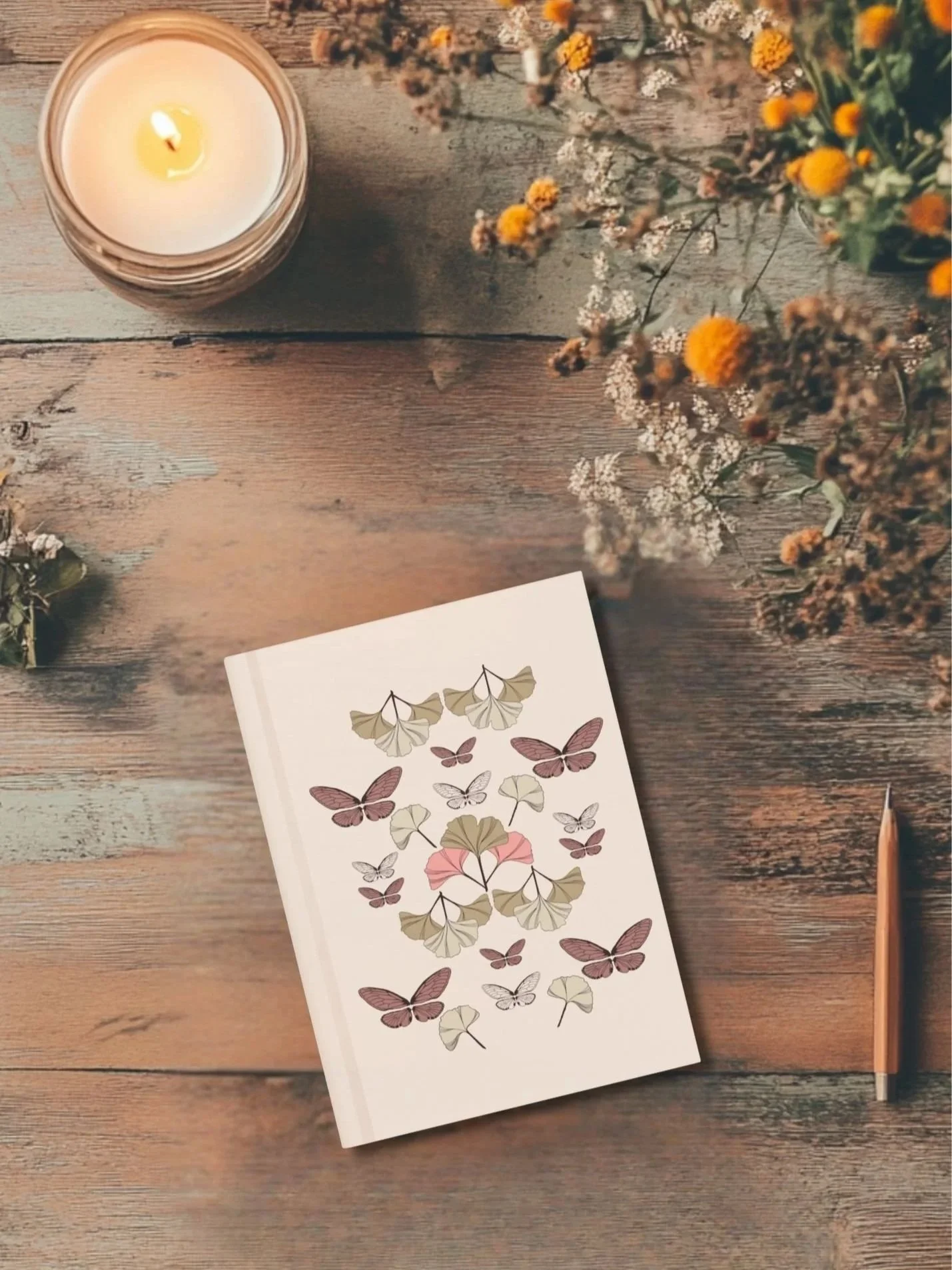 Botanical journal with ginkgo leaves and butterfly design placed on wooden surface beside candle and yellow floral arrangement- warm and cozy workspace.