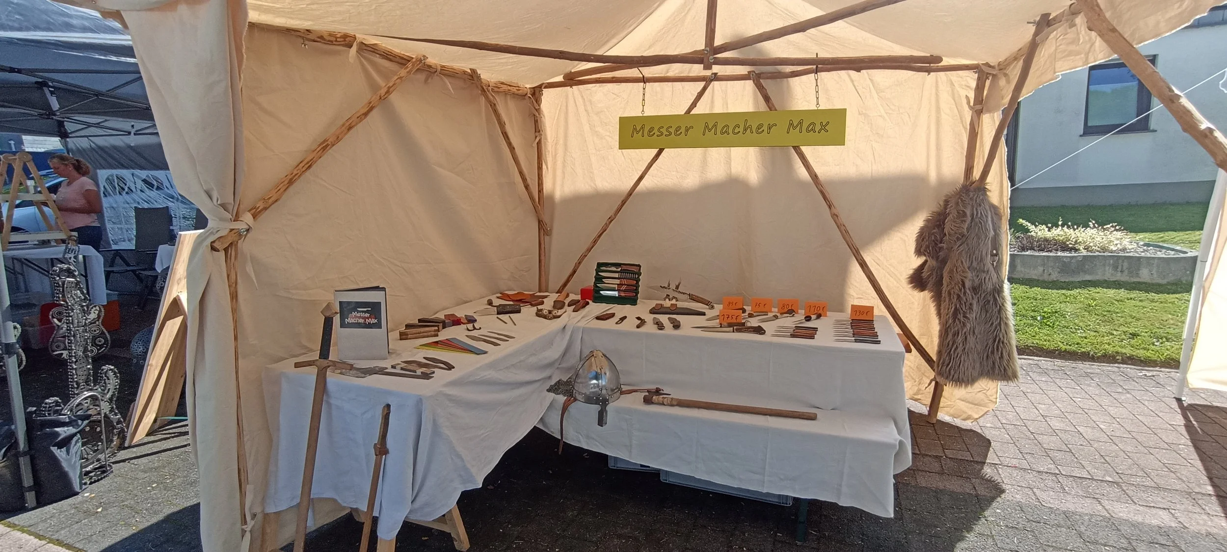 Stand mit Holzwerkzeugen und Zubehör bei einem Markt, mit einem Schild 'Messer Macher Max', Holzwaffen und einem Fell auf der rechten Seite.