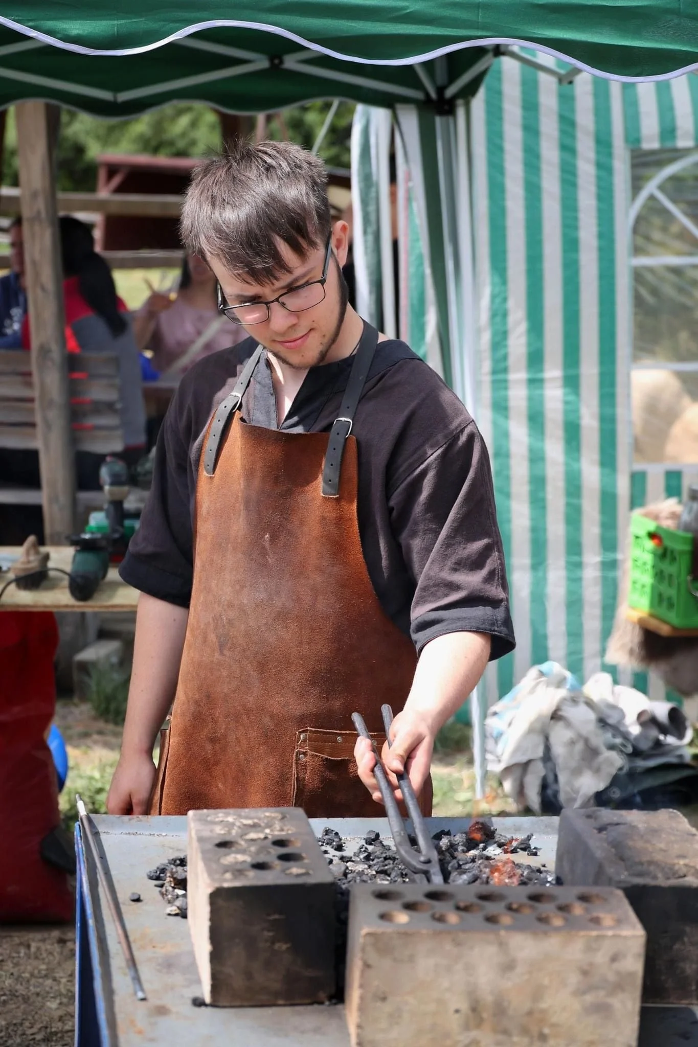 Junger Mann in Lederjacke und Brille, der auf einem Grill Feuer macht, im Freien bei einer Veranstaltung.