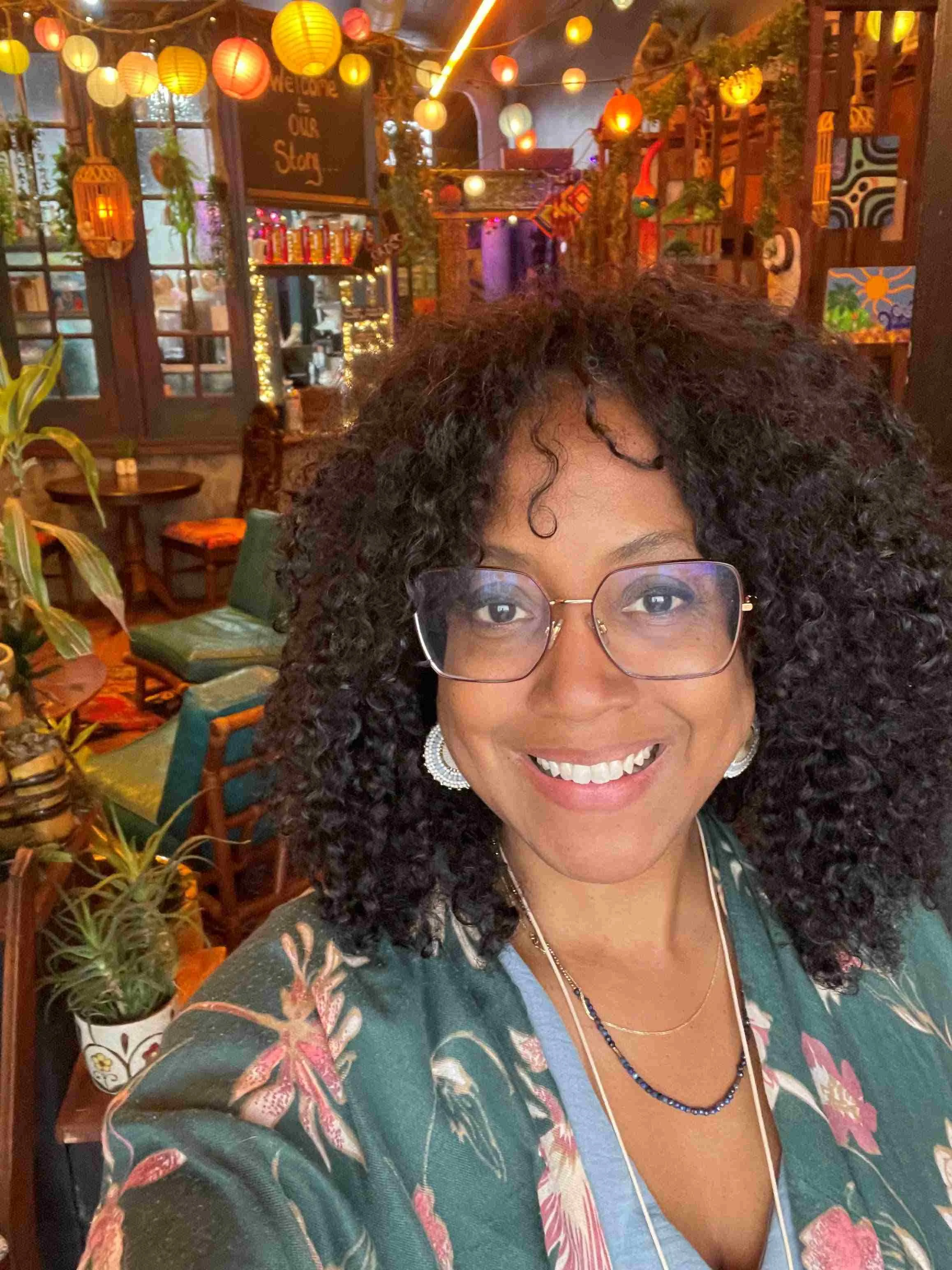 A woman with curly black hair and glasses smiling at the camera in a vibrantly decorated indoor setting with hanging colorful lanterns and wall art.