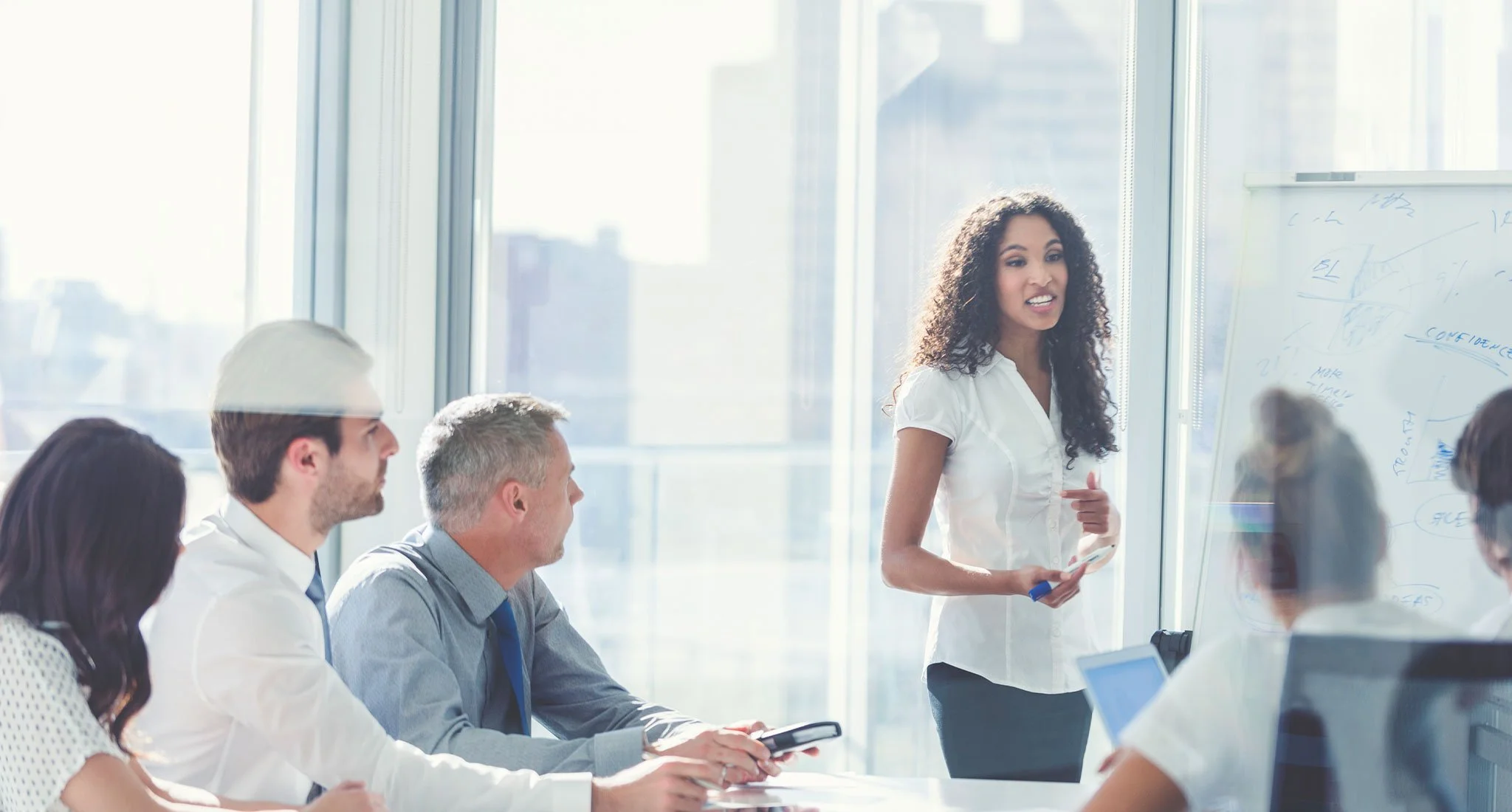 A woman presenting to a group in a modern office with large windows, whiteboard, and city view.