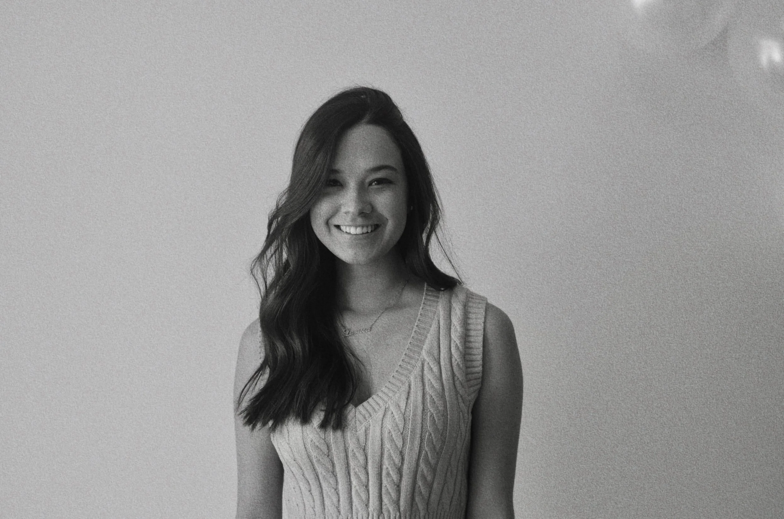 Black and white photograph of a young woman smiling, with long wavy hair, wearing a sleeveless knitted top and a necklace, standing against a plain background.