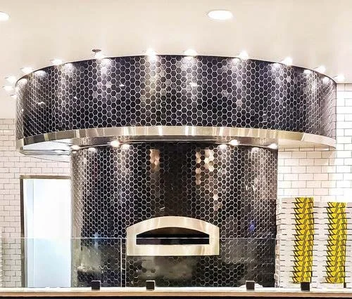 Modern pizza oven with black hexagonal tiles and a stainless steel arch, situated in a contemporary restaurant setting with white subway til
