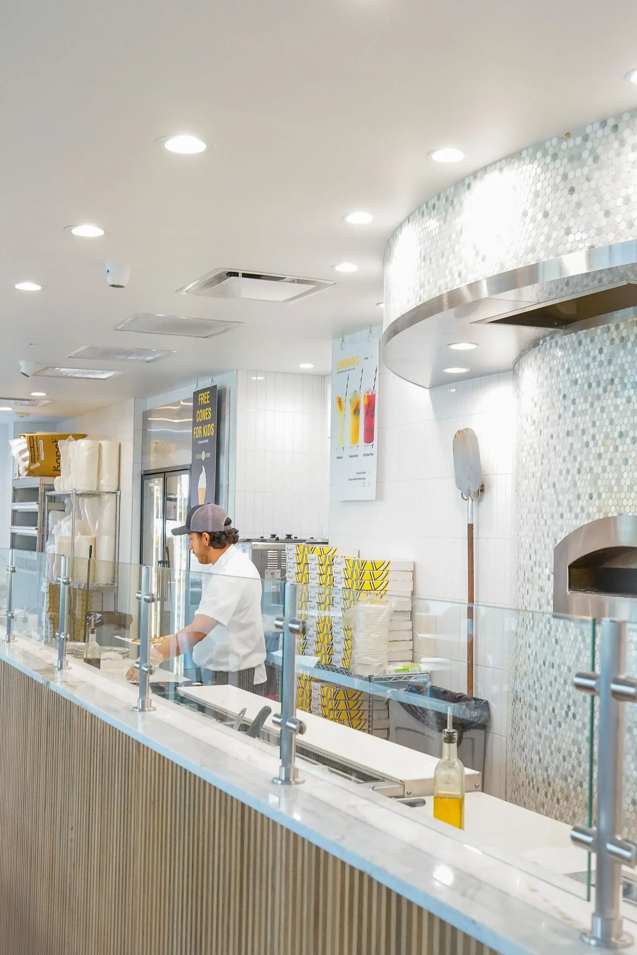 Inside a modern ice cream shop with a worker preparing food behind a white marble counter. The background includes colorful signage and a tiled wall.